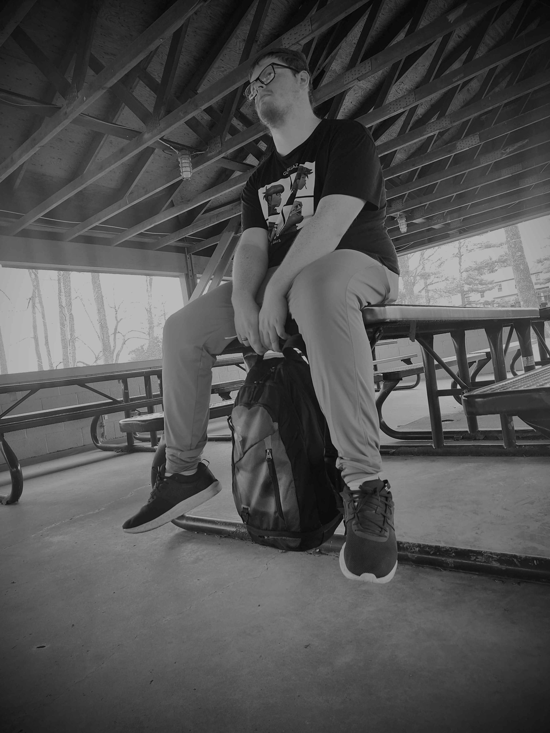 Young person sitting on a backpack under a wooden pavilion, with picnic tables and trees in the background.