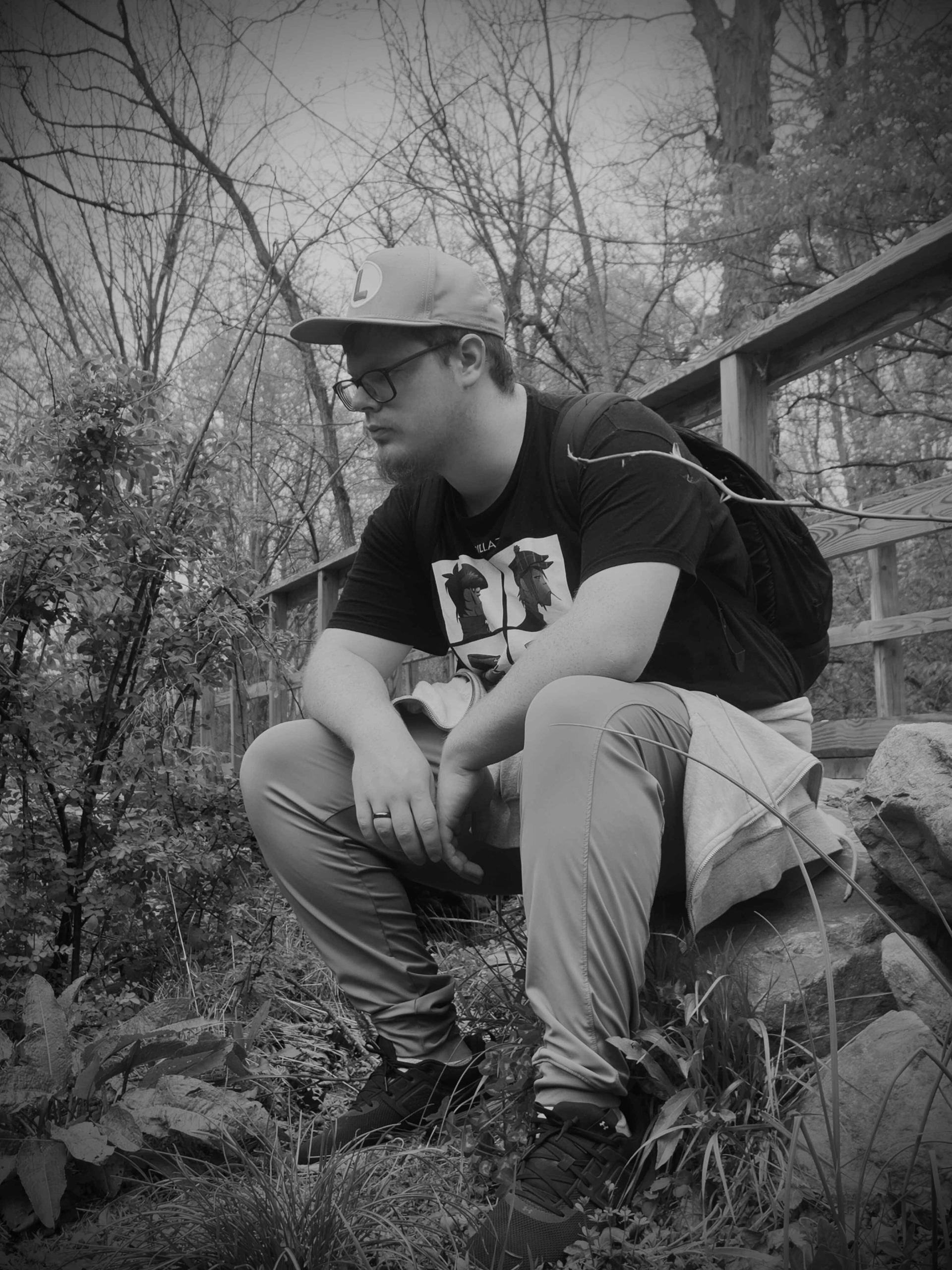 Person sitting outdoors on rocks, wearing a cap, glasses, and casual clothing, surrounded by trees and foliage.