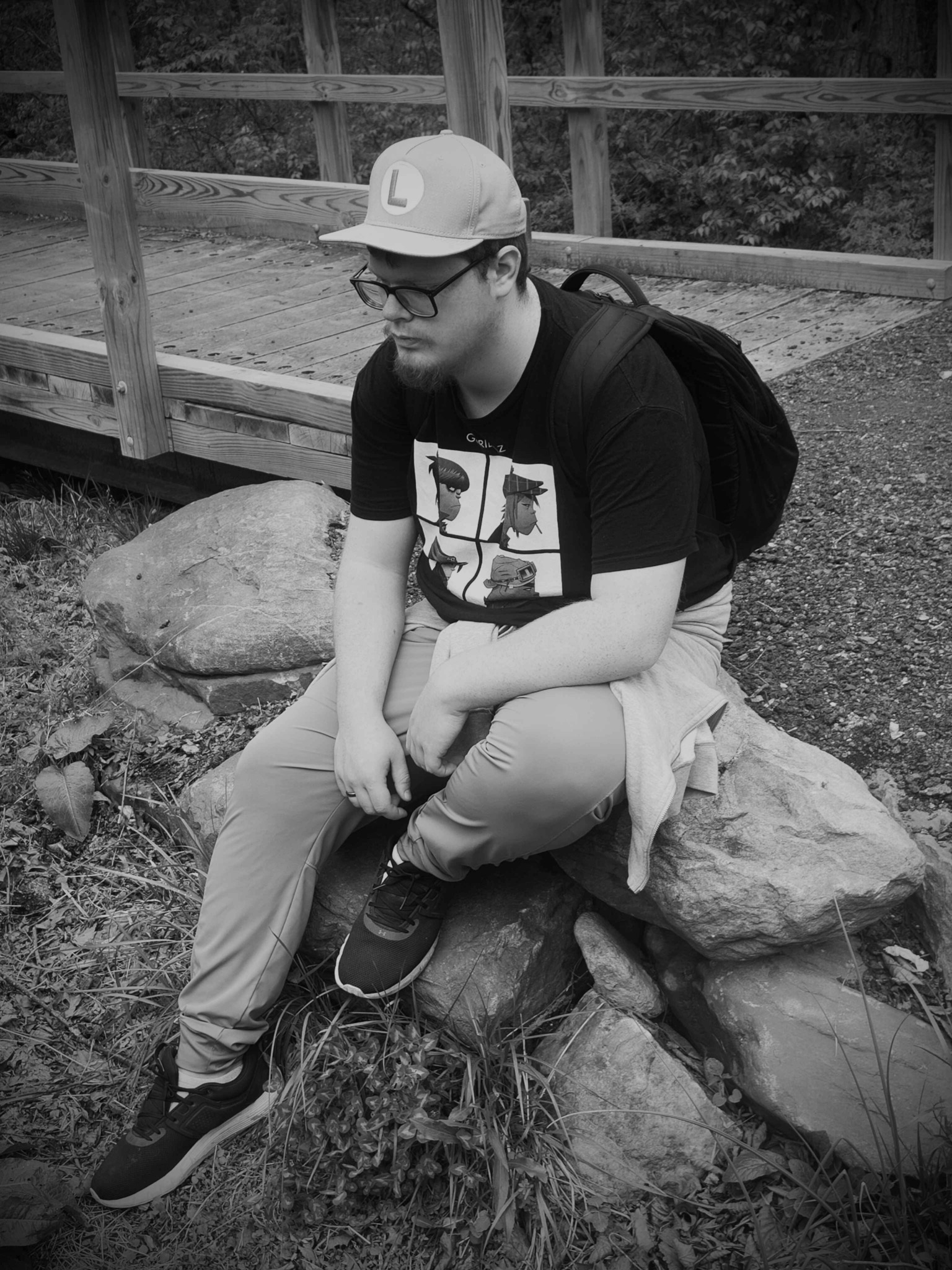 Person sitting on a rock outdoors, wearing glasses, a cap, and a backpack, with a wooden fence in the background.