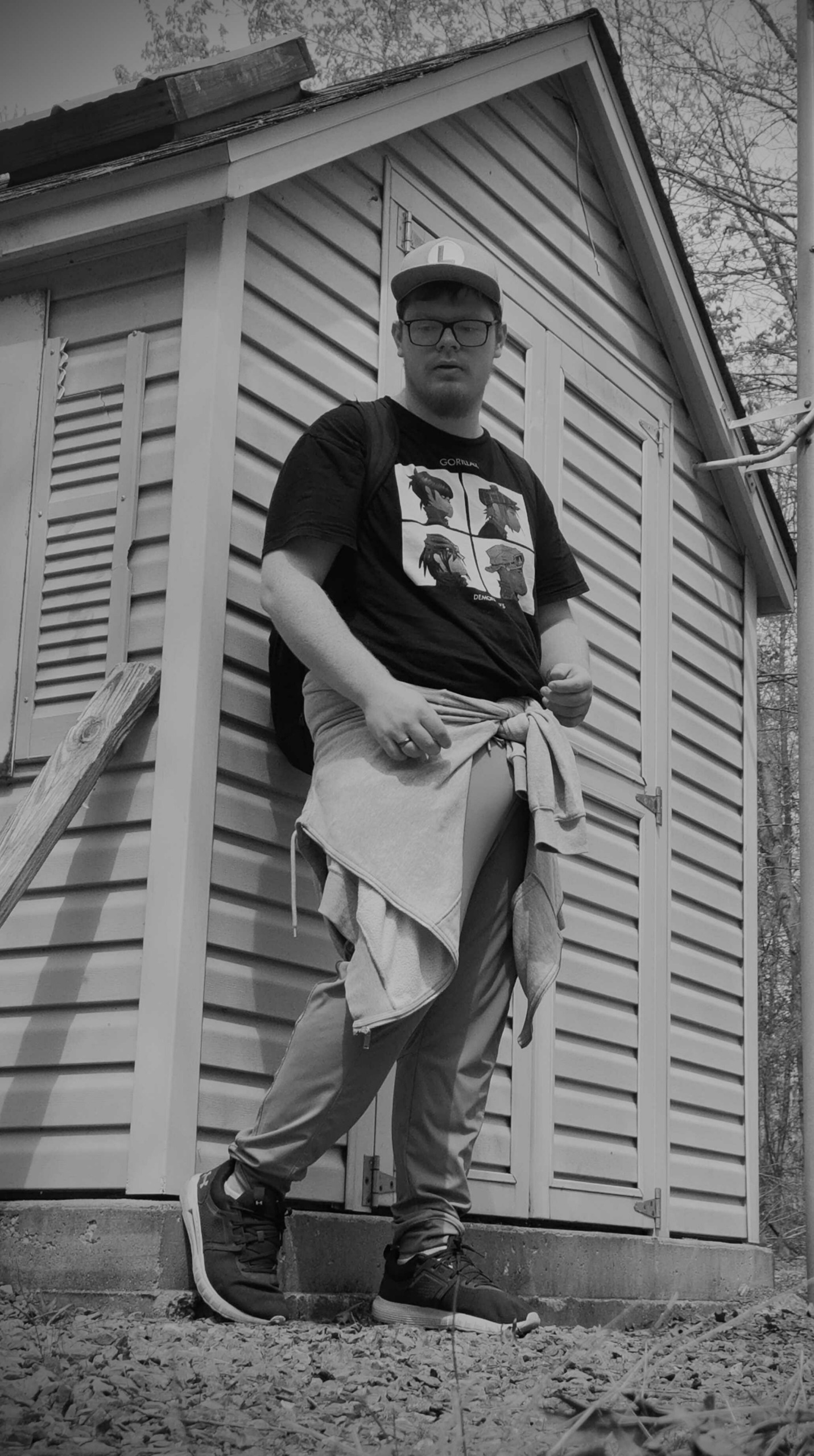 Person standing outside a house with siding, wearing a cap, glasses, a graphic t-shirt, and pants, holding a jacket.