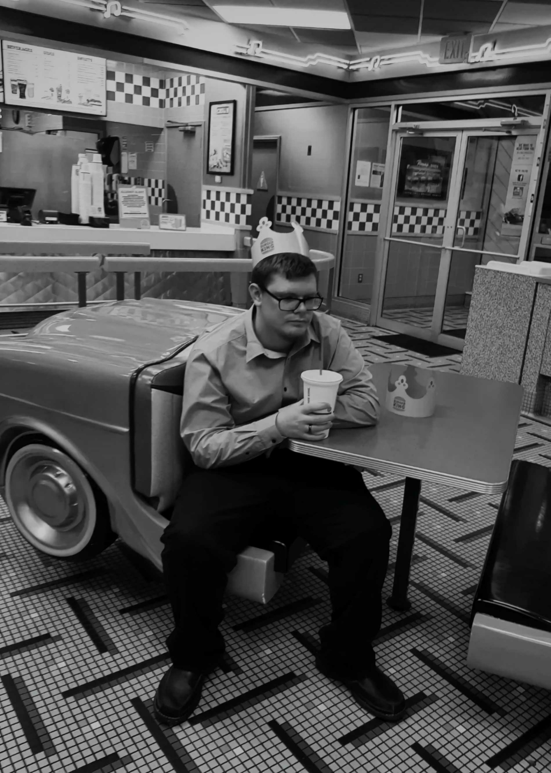 Man sitting at table in fast food restaurant, holding drink, with checkered decor and counter in background.