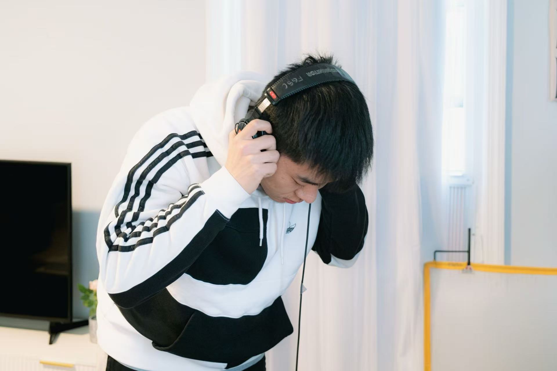 Young person wearing headphones, leaning forward, in a bright room with curtains and a yellow object in the background.
