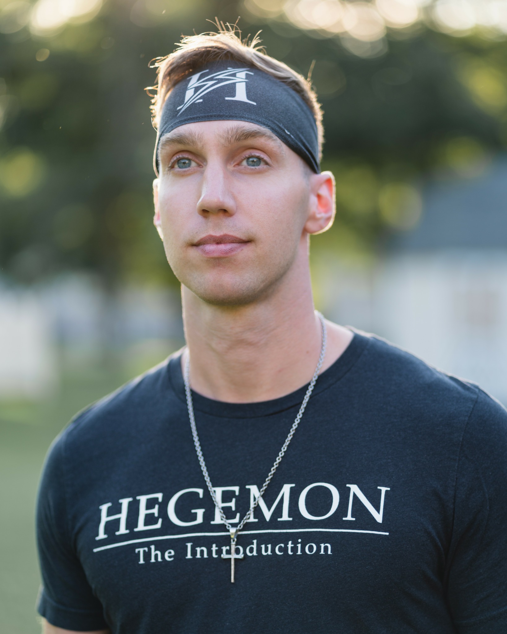 Young man with headband and necklace outdoors, wearing a black T-shirt with text, blurred background of trees and sky.