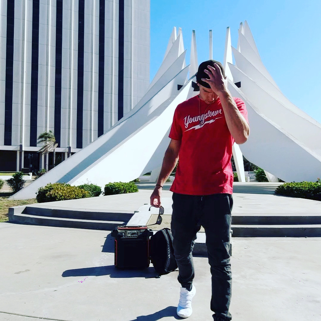 Person standing outdoors in front of a modern white architectural structure, holding a phone to their face, with a suitcase nearby.
