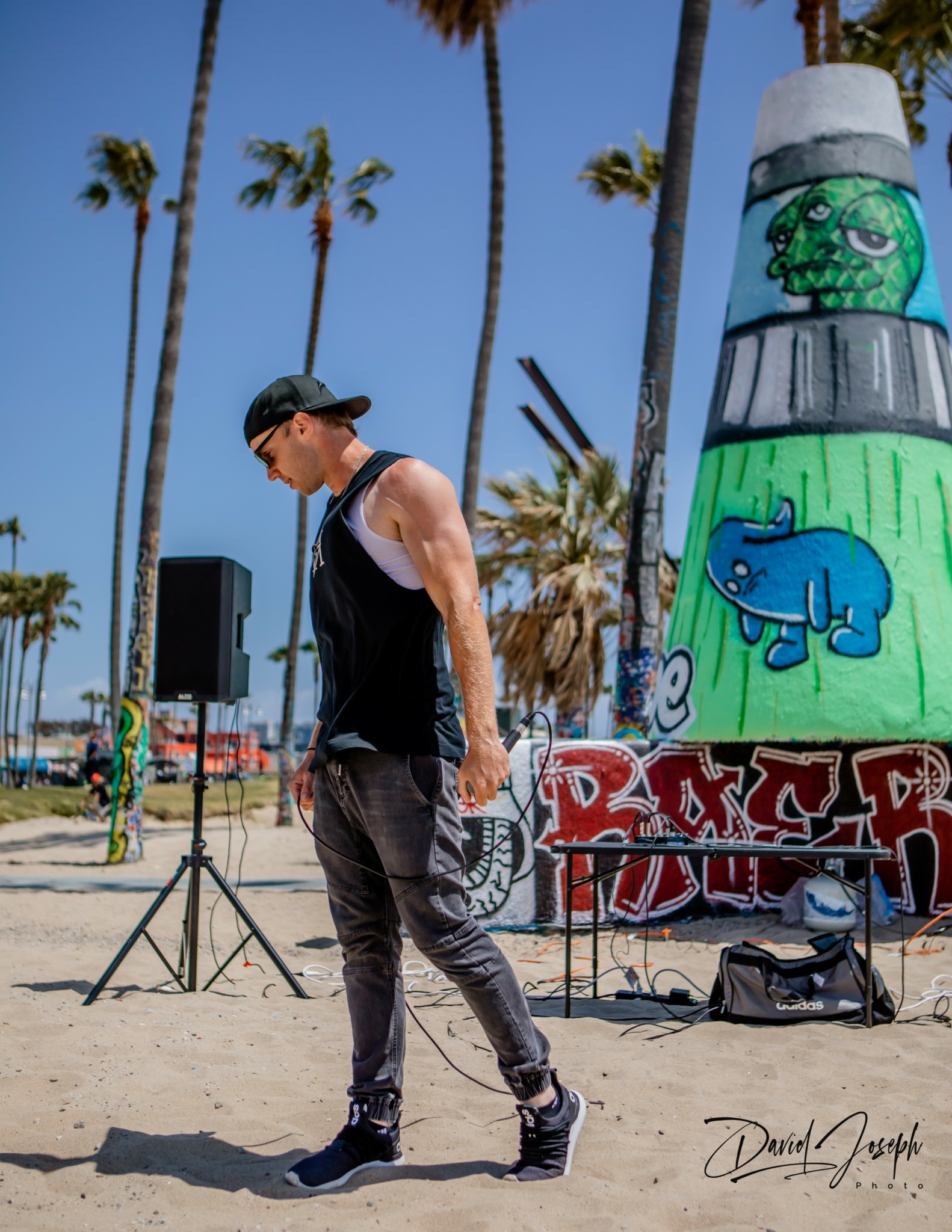 Young man walking on beach with palm trees and graffiti-covered lighthouse in background.