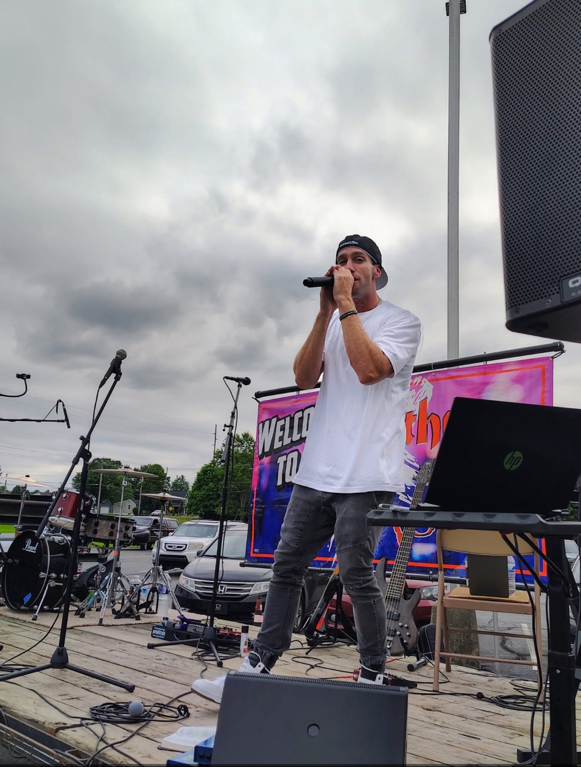 Person singing into microphone on outdoor stage with equipment and cloudy sky background.