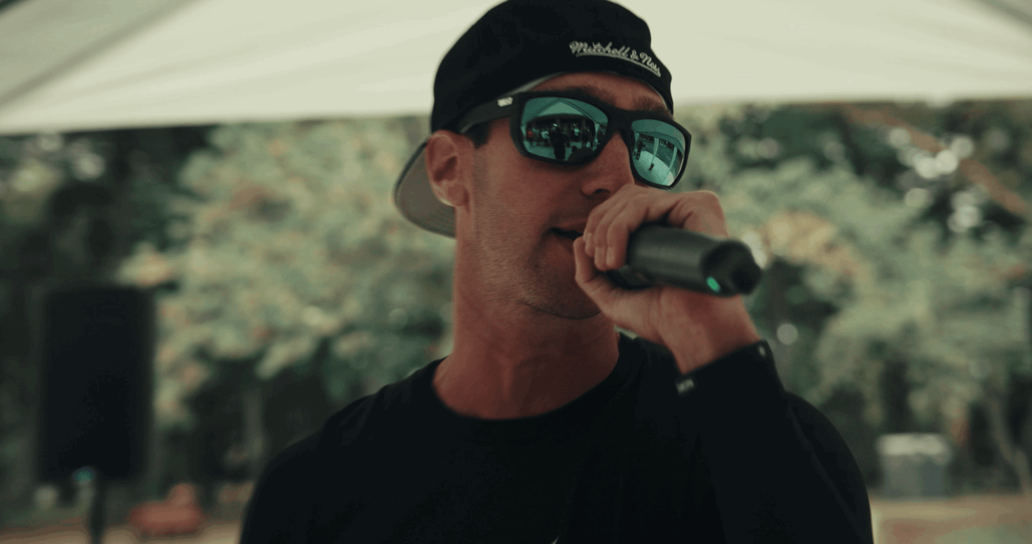 Person wearing sunglasses and a cap, holding a microphone outdoors with trees in background.