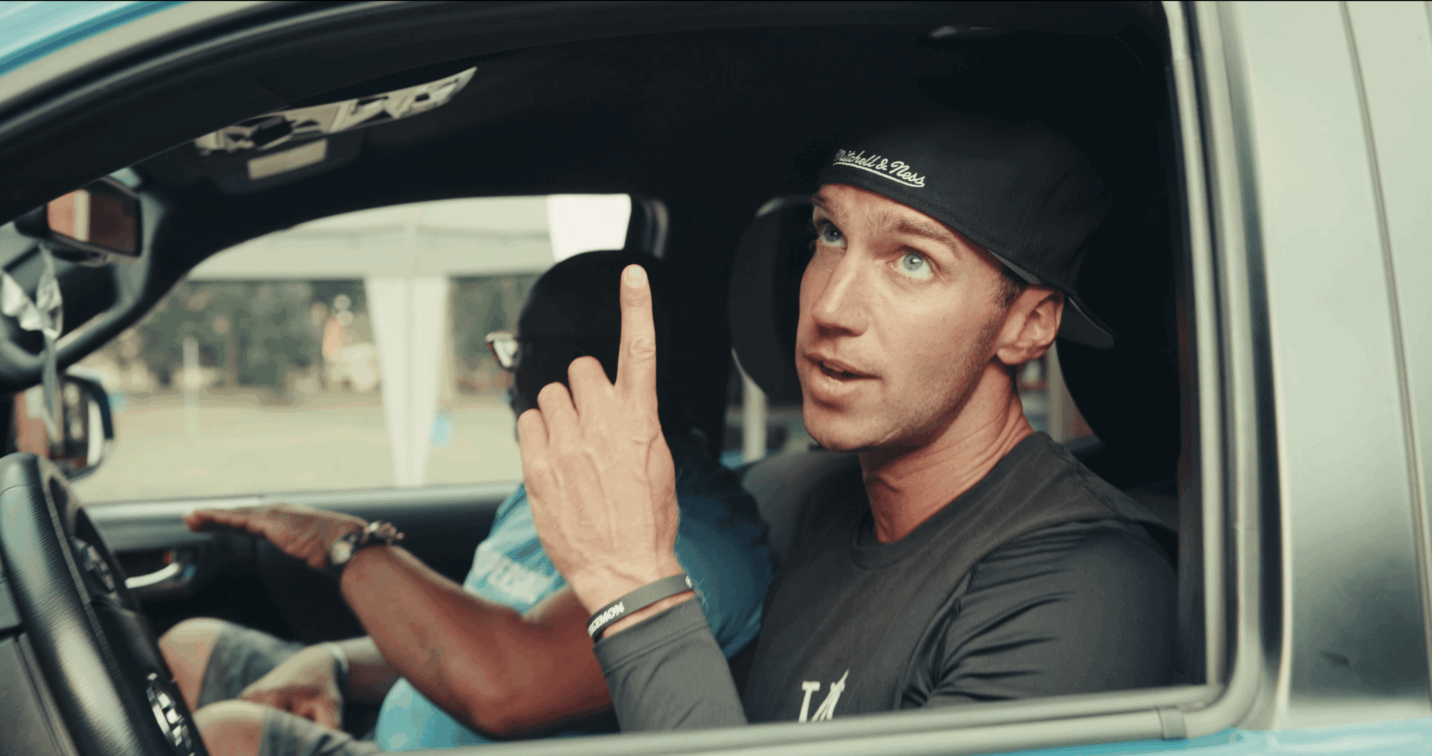 Man wearing a black cap and shirt, sitting in a vehicle, gesturing with his index finger, speaking.