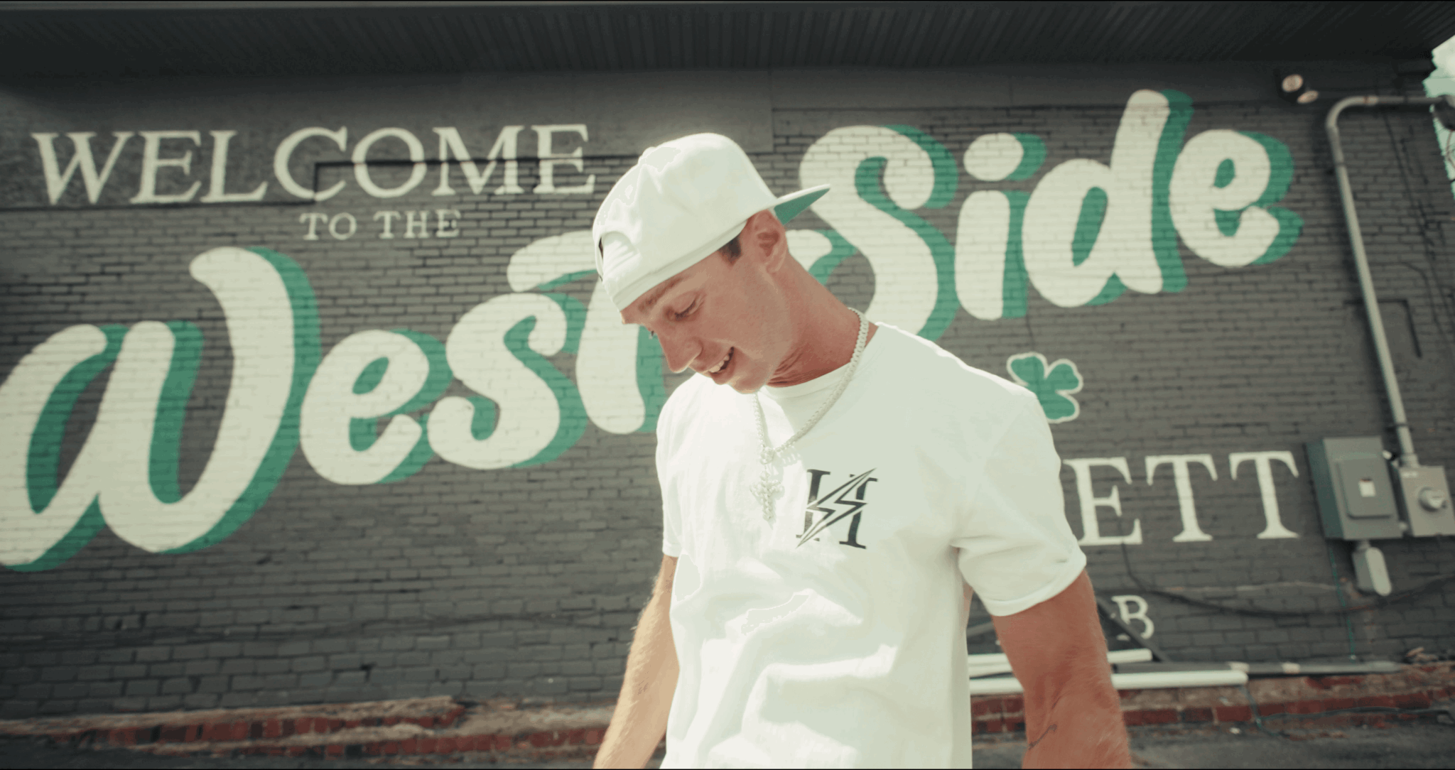 Young man wearing a white hat and t-shirt standing in front of a mural that says 'Welcome to the West Side'.