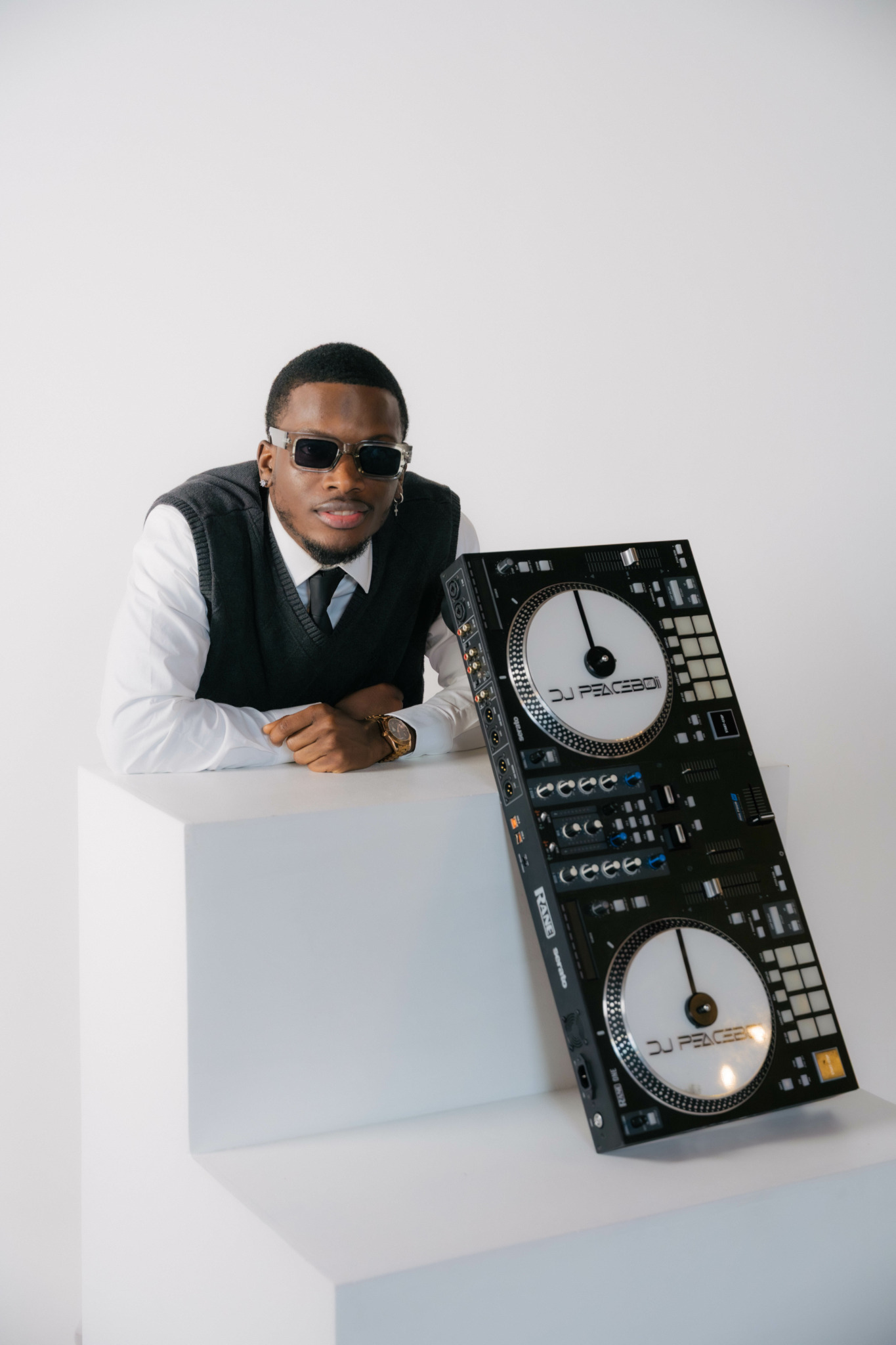 Man in sunglasses and formal attire leaning on a white surface with DJ equipment beside him.