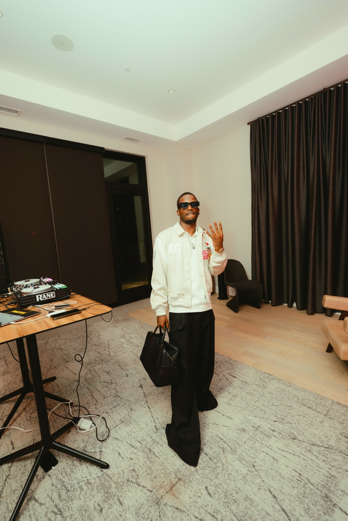 Person wearing sunglasses and white jacket holding a black bag, standing in a room with dark curtains and wooden floor.