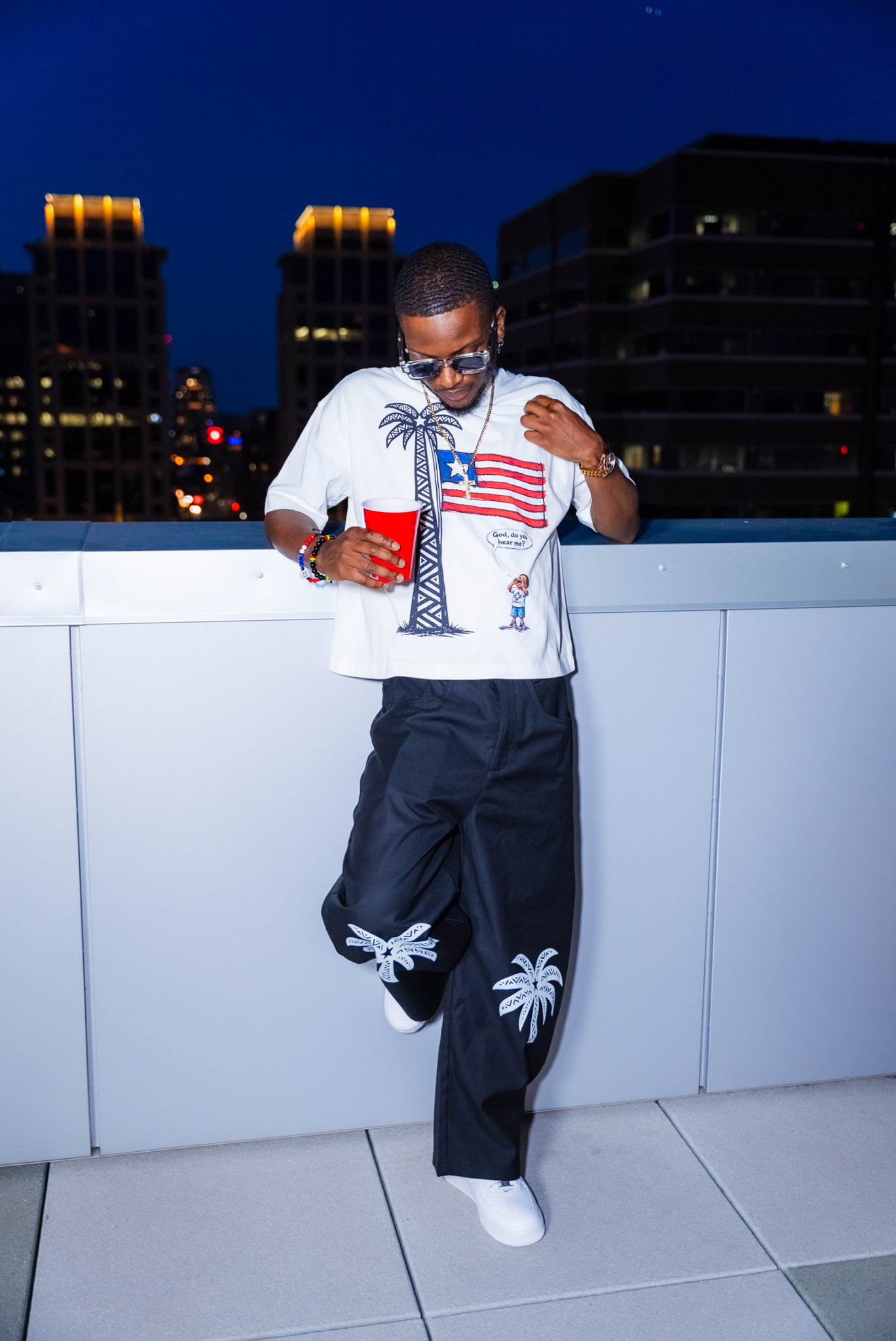 Young man in sunglasses and casual clothing leaning against a white railing at night, holding a red drink.