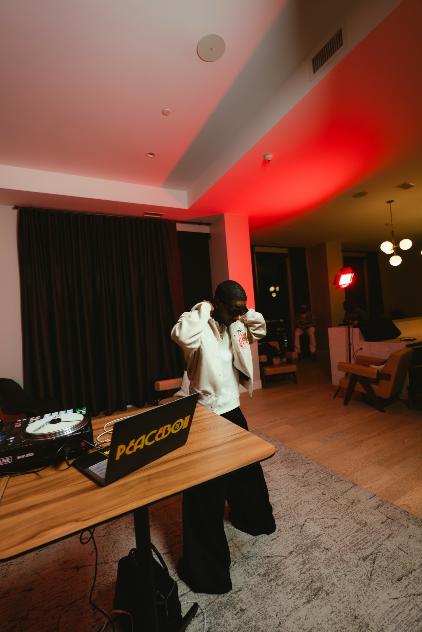 Person dancing indoors with a DJ setup on a table, dark curtains, and warm lighting in the background.