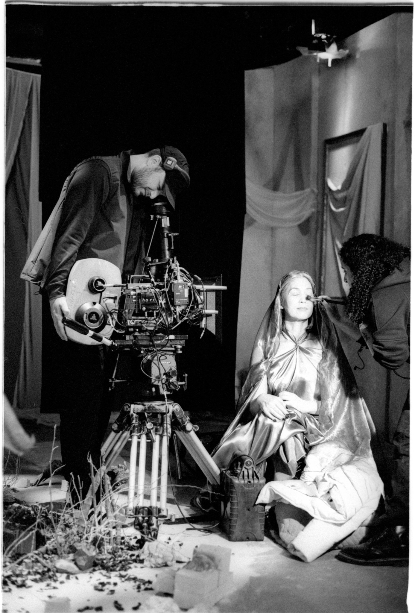 Person operating camera equipment with a woman sitting on the floor and another person adjusting her hair or makeup.