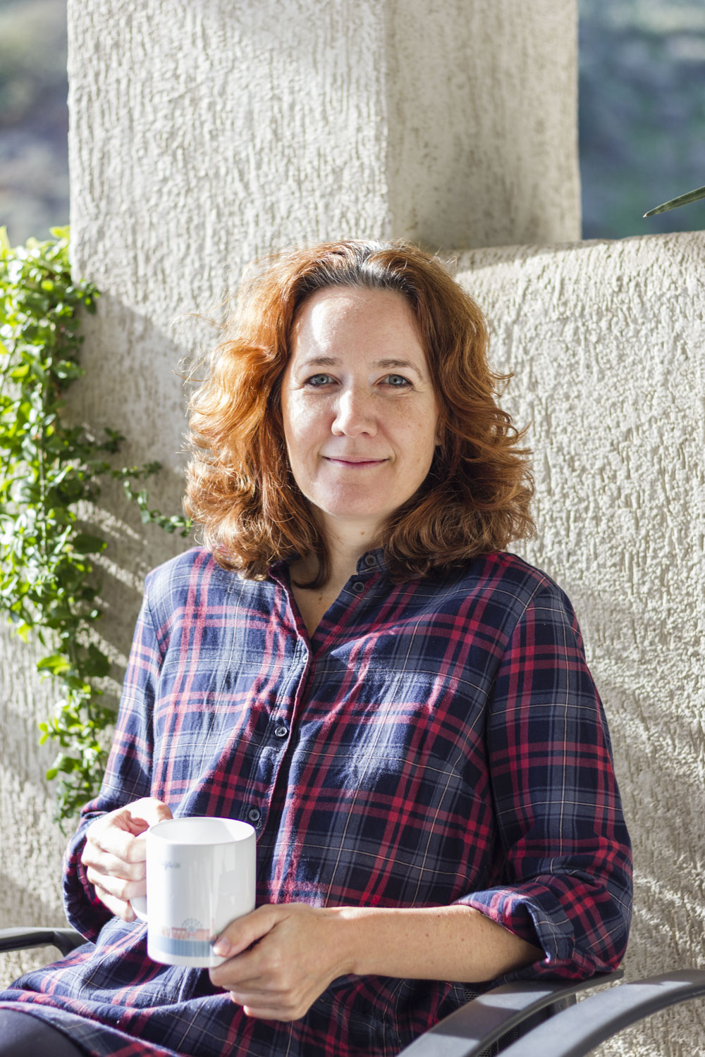 Woman with curly hair holding a mug, standing outdoors near a stone wall and greenery.