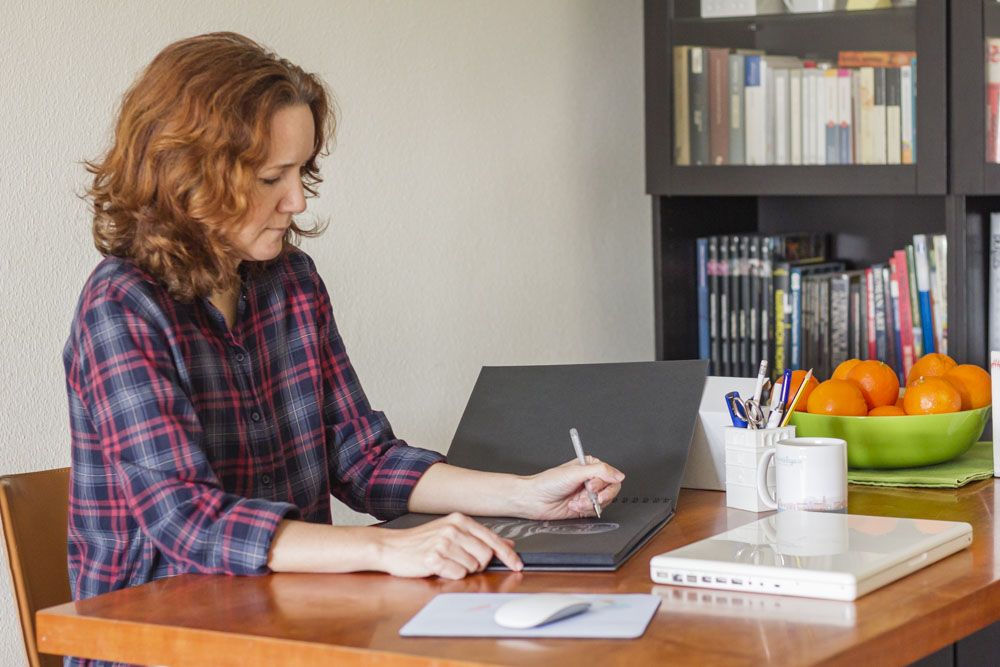 Woman with curly red hair in plaid shirt working at desk with laptop and papers, bookshelf with books and fruit bowl nearby.