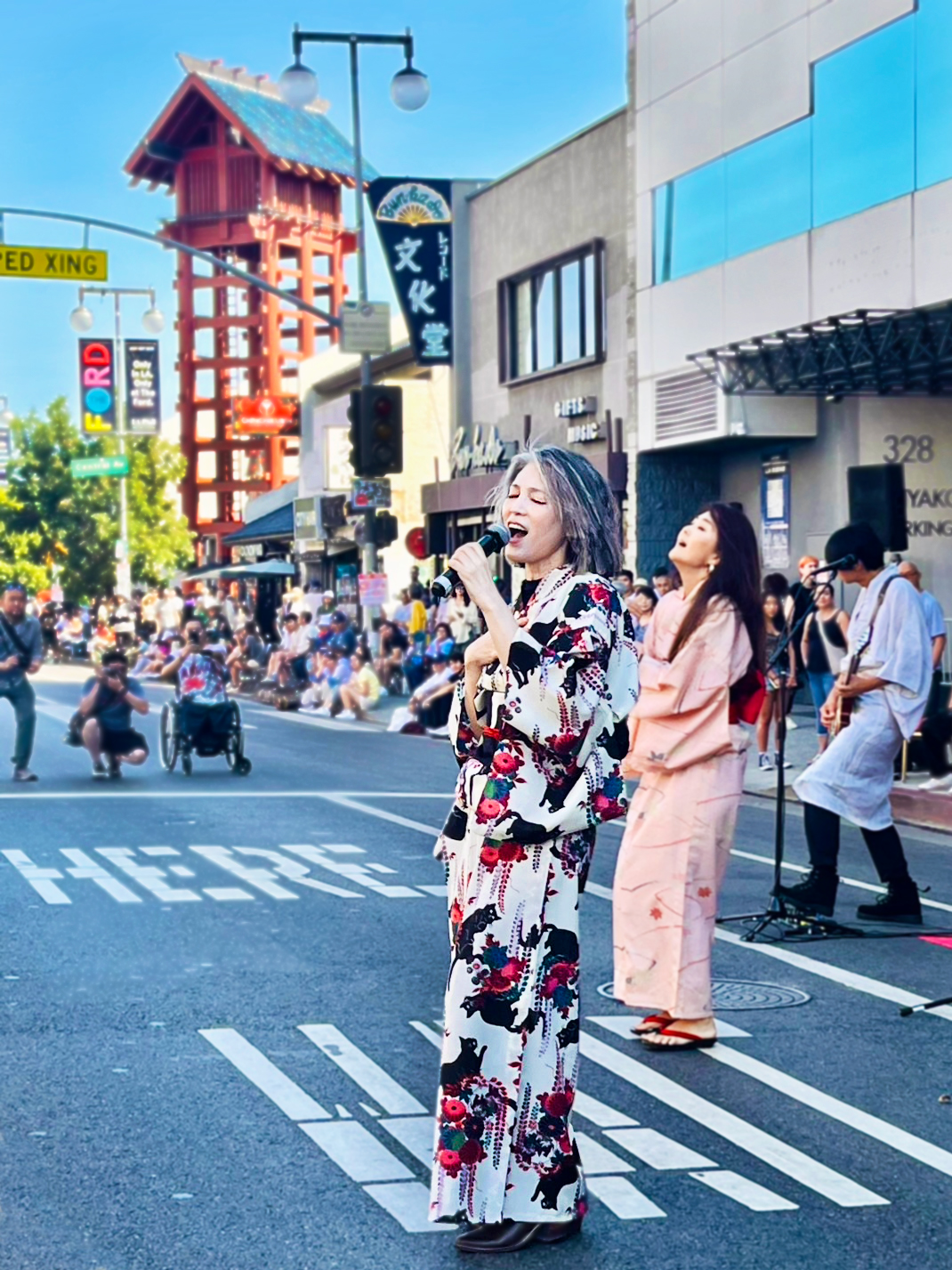 Woman in floral yukata standing on crosswalk holding a phone, busy city street with pedestrians and buildings.