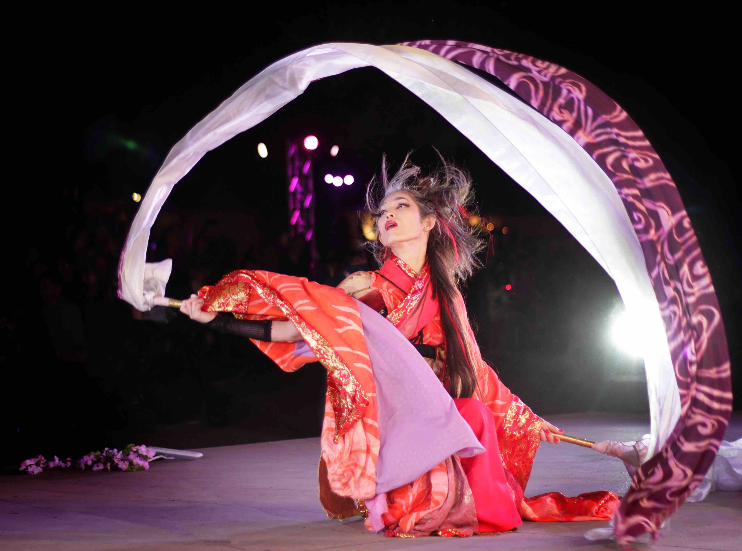 Person in traditional costume performing with flowing fabric on stage at night.