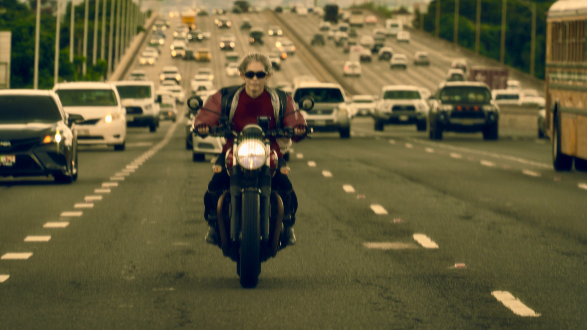 Person riding a motorcycle on a busy multi-lane highway with cars and buses, wearing sunglasses and a jacket.