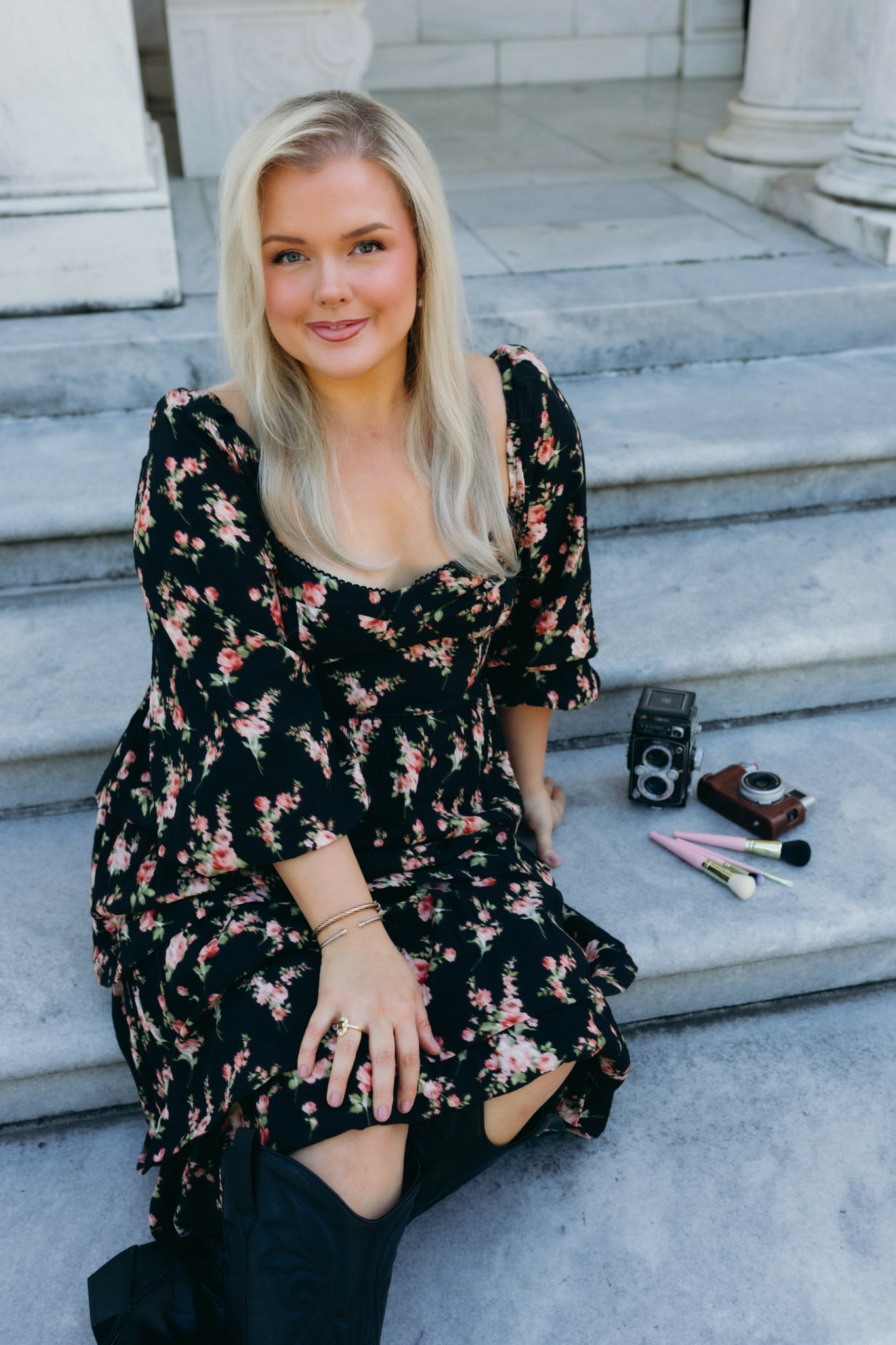 Young woman with long blonde hair sitting on stone steps, wearing a floral dress, smiling, with makeup and accessories.