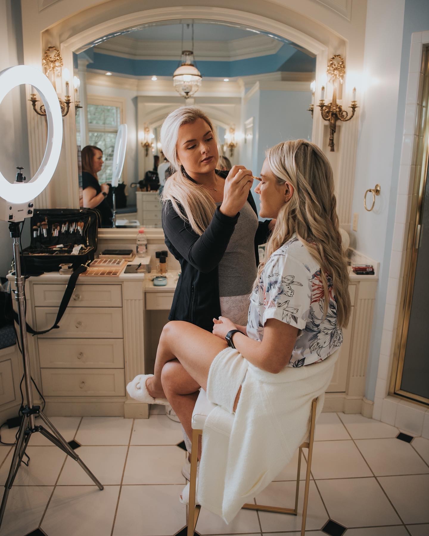Makeup artist applies makeup to a woman seated on a chair in a well-lit room with elegant decor.