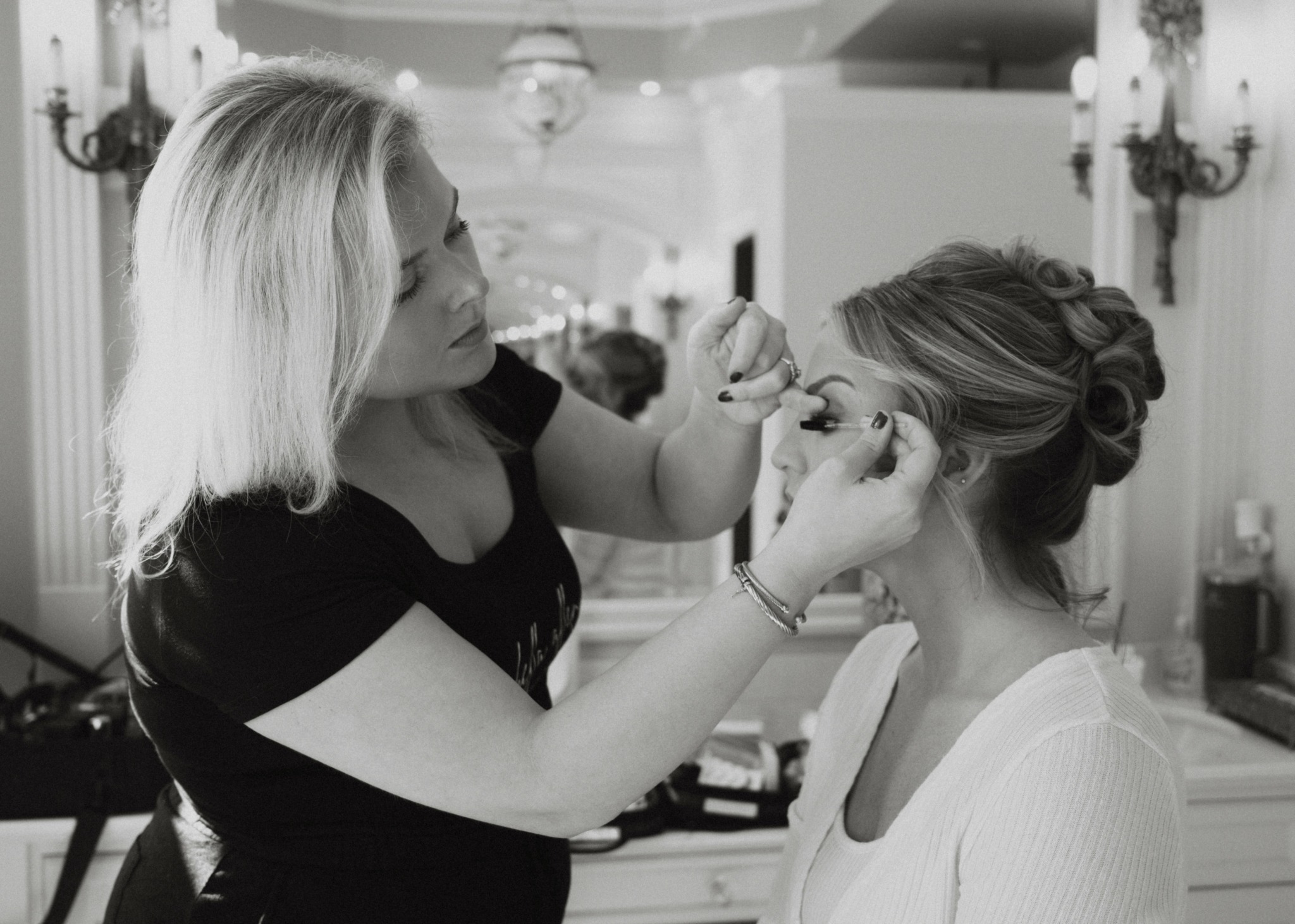 Makeup artist applies makeup to a seated woman in a room with elegant decor.