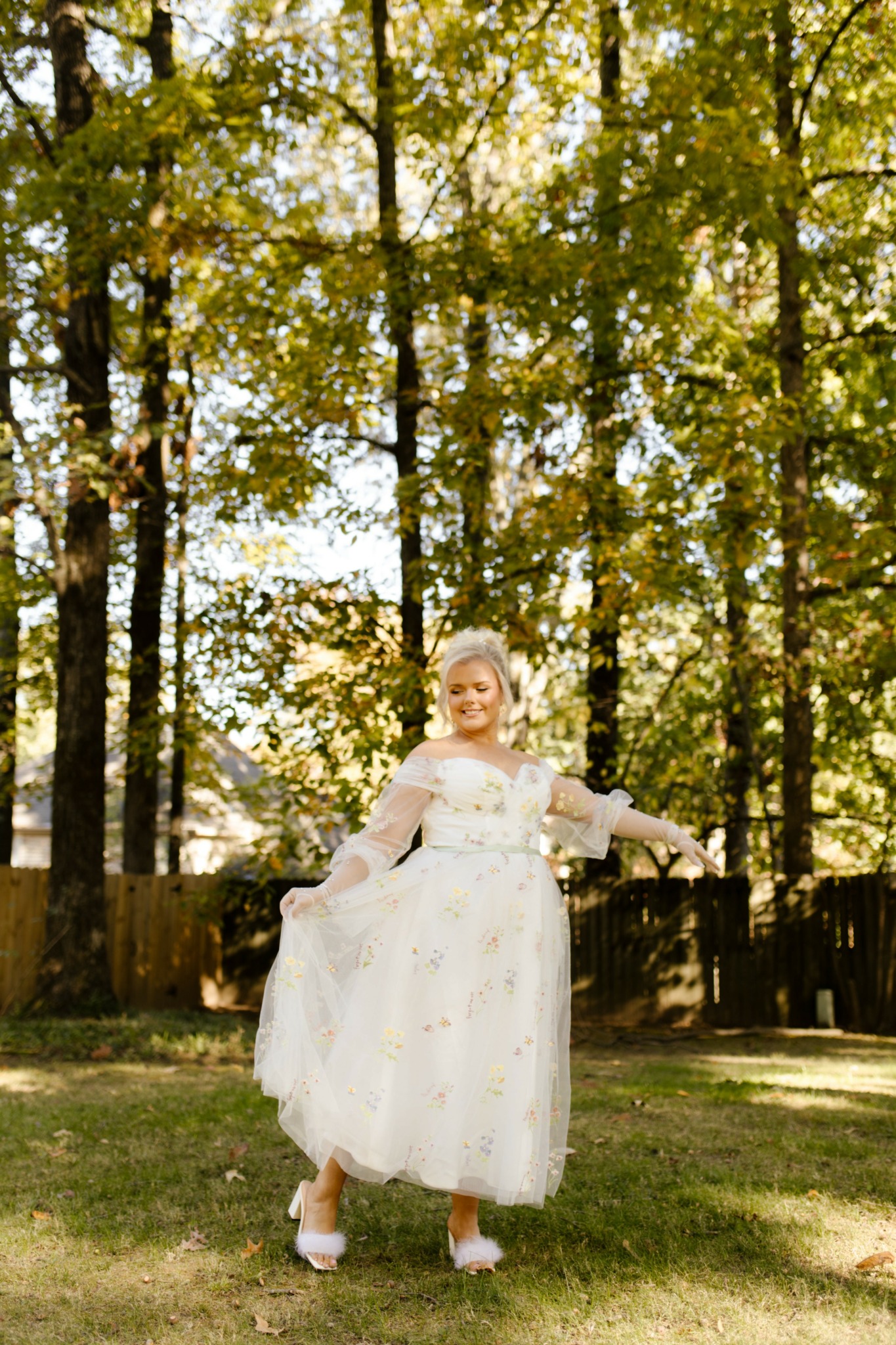 Woman in a white dress standing outdoors among trees with yellow and green leaves, smiling and holding her dress.