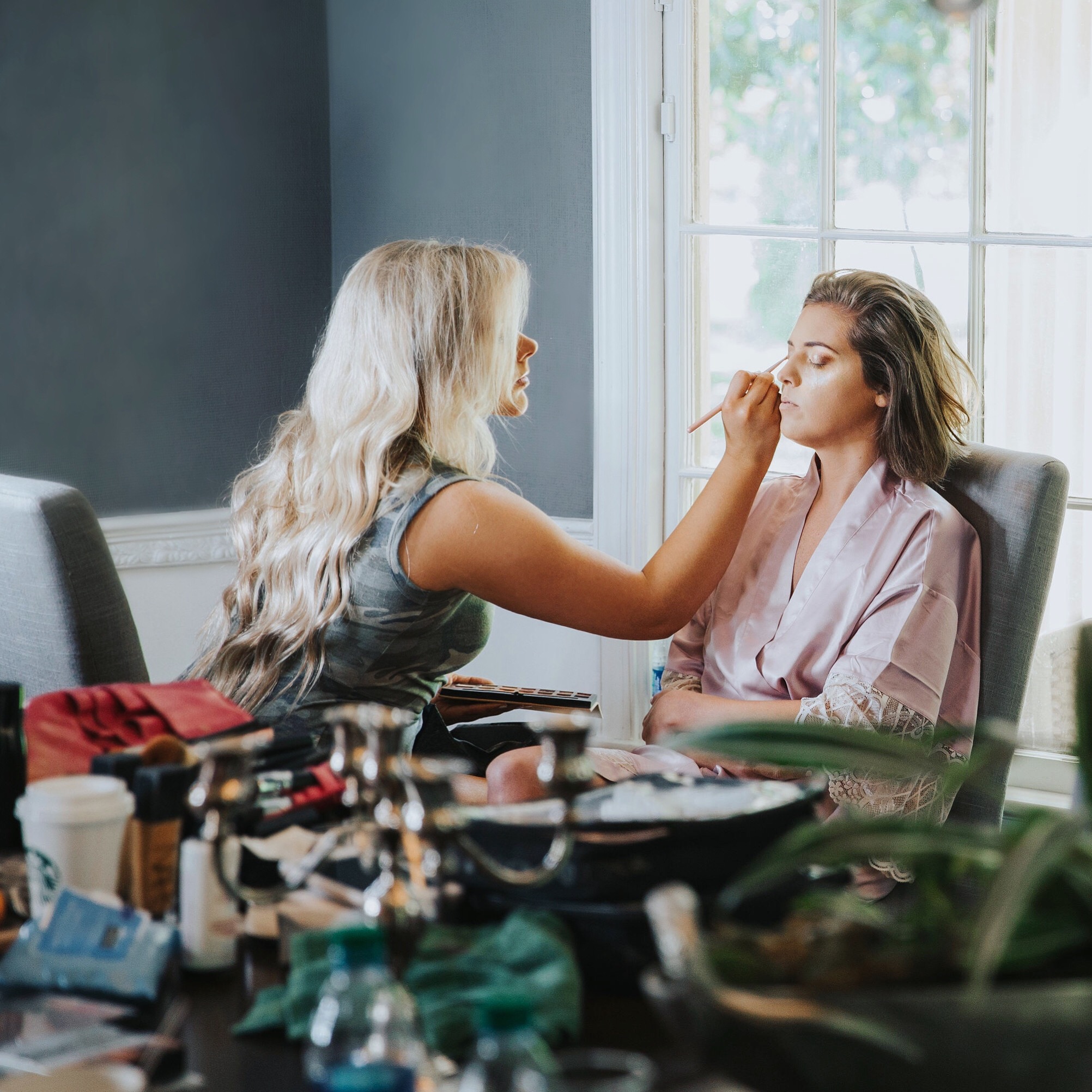 Makeup artist applying makeup to a woman sitting by a window in a room with a table full of makeup products.
