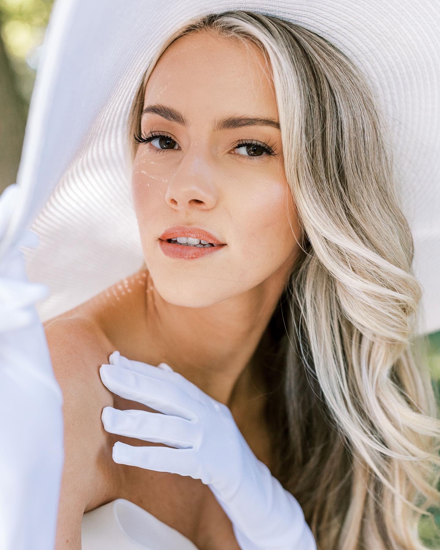 Young woman with long, wavy blonde hair, wearing a white glove and holding a white umbrella outdoors.