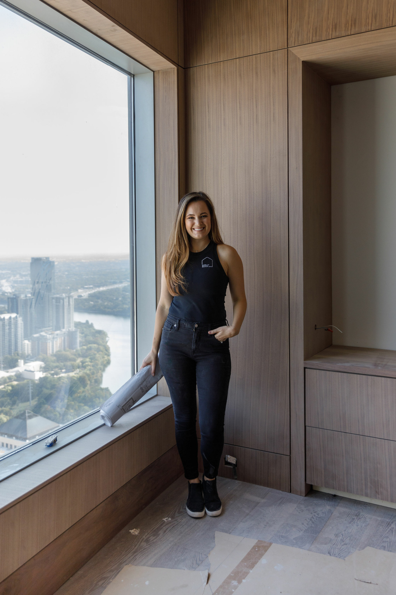 Young woman with long hair standing by a large window overlooking a cityscape, smiling, wearing a dark sleeveless top and jeans.