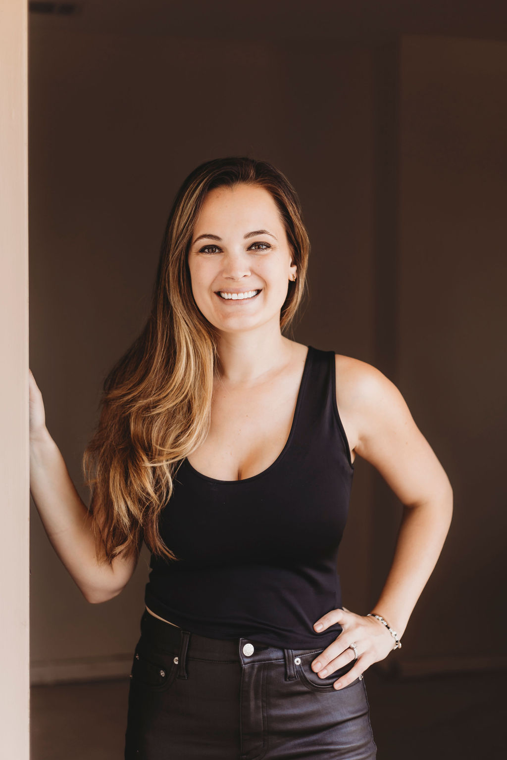 Woman with long hair smiling, wearing a black tank top and dark pants, standing indoors.