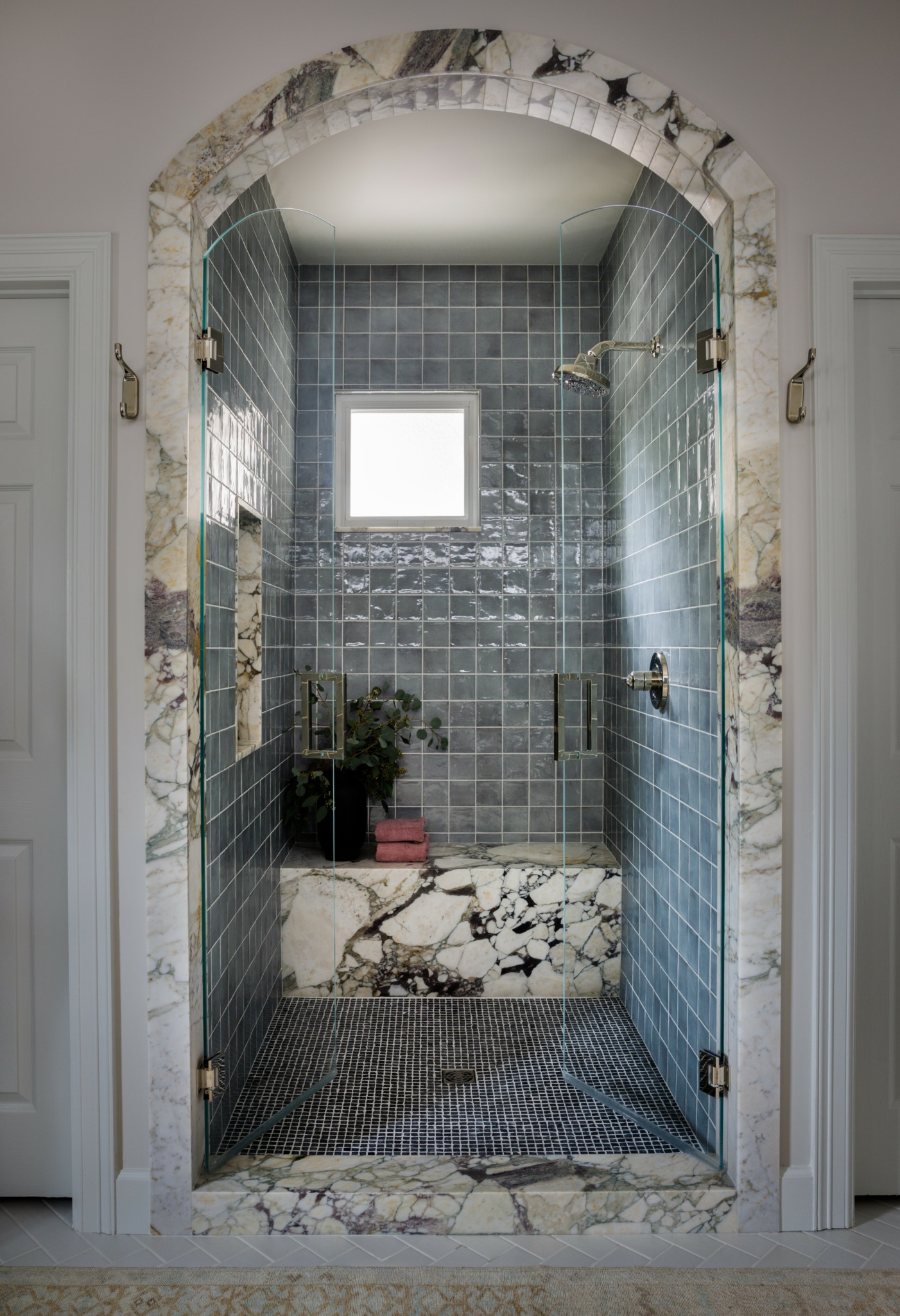 Shower area with blue tiled walls, small window, and marble ledge, viewed through an arched doorway with stone trim.