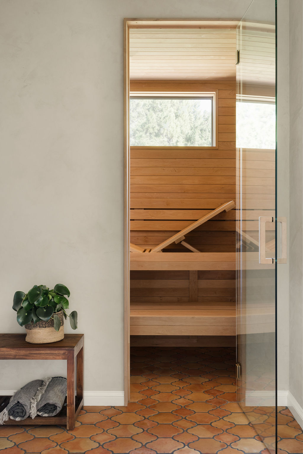 Part of a room with a wooden sauna visible through a glass door, a small wooden table with a plant, and a patterned tile floor.