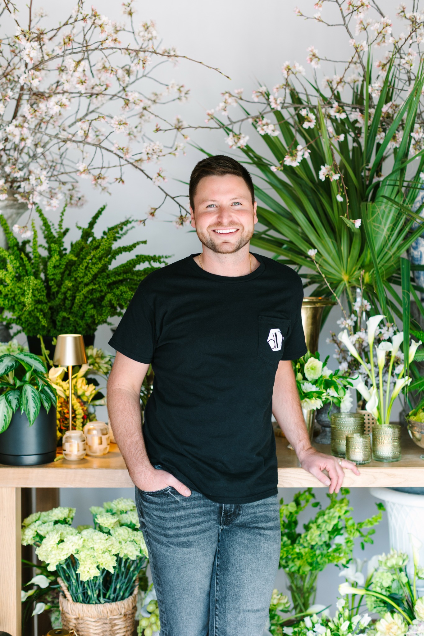 Man with short dark hair and beard smiling, standing in front of a lush plant display, wearing a black t-shirt and jeans.