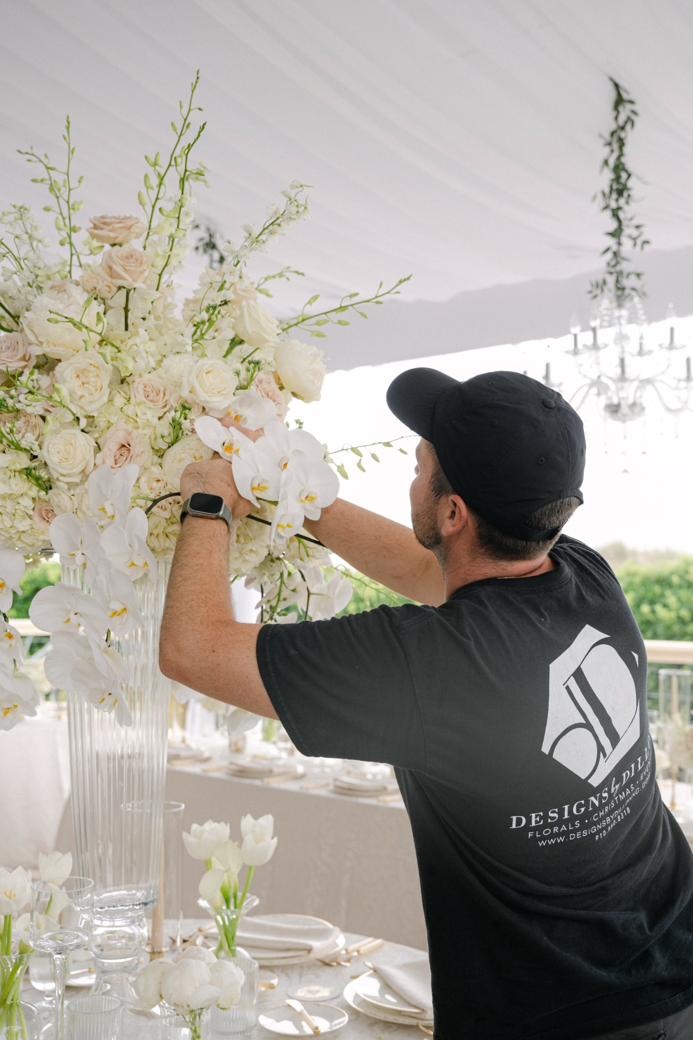 Person arranging a large floral centerpiece with white and pink flowers at a table setting.
