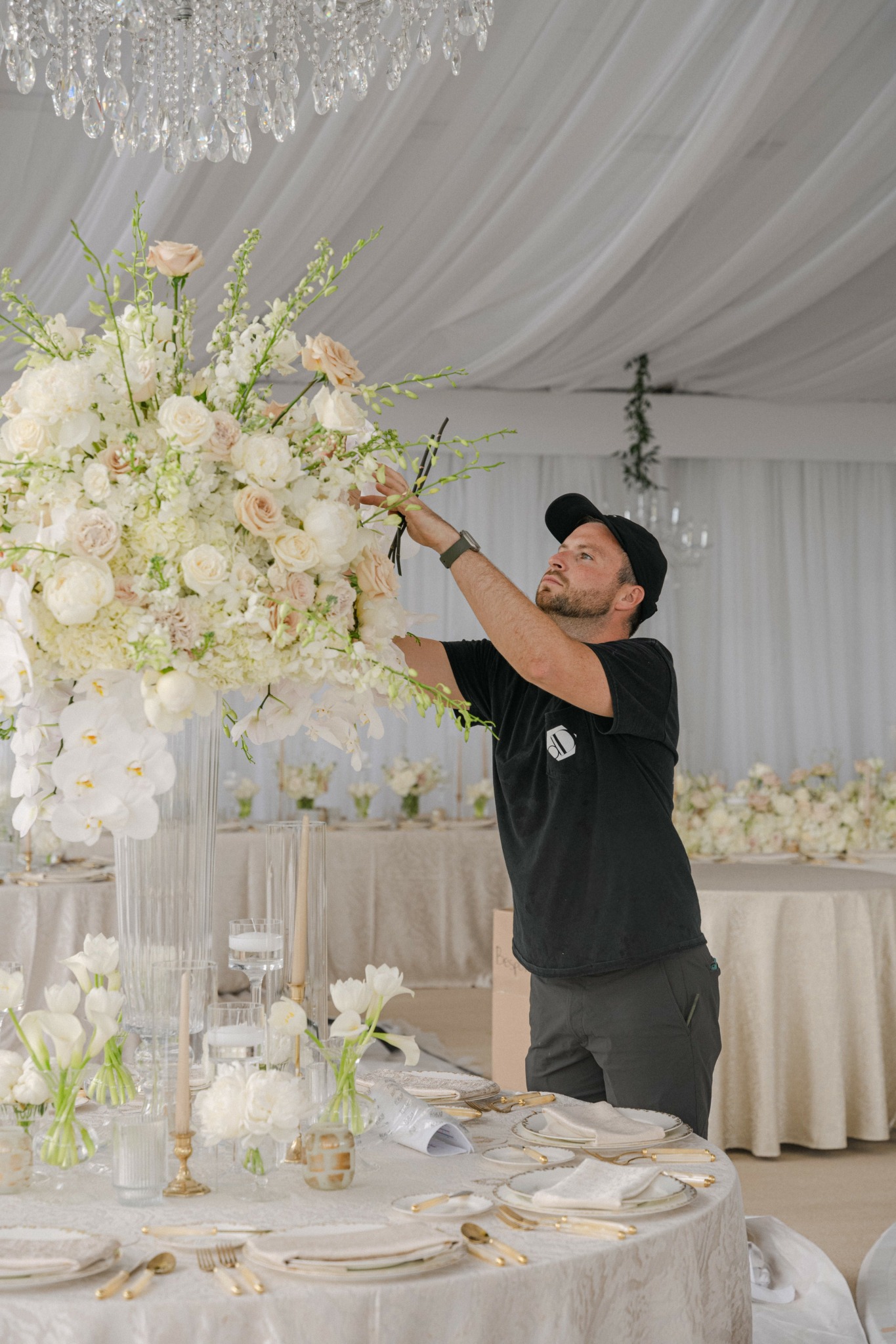 Man arranging large floral centerpiece at a decorated event table with white flowers and elegant tableware.