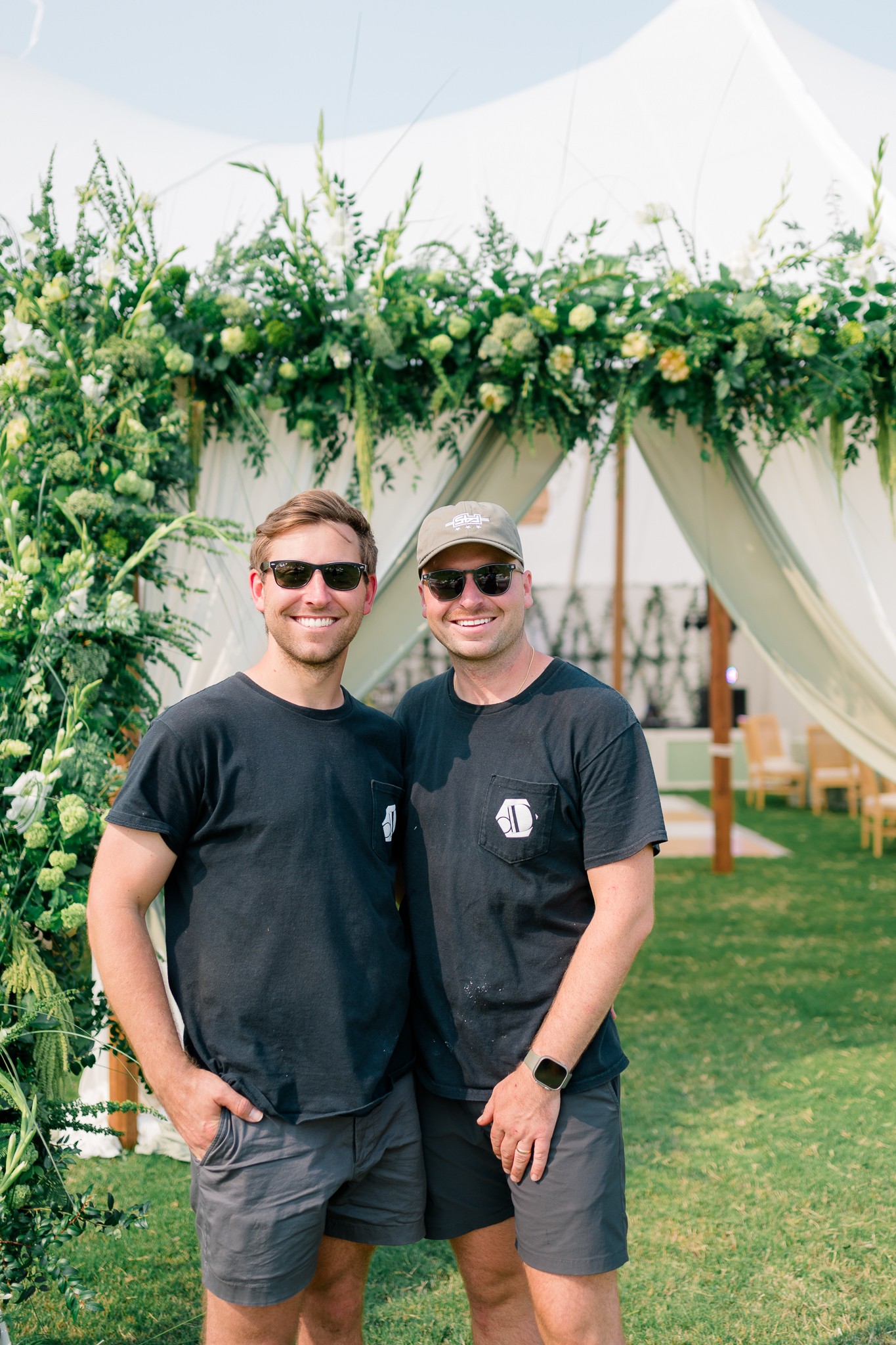 Two men stand outdoors in front of a floral arch, smiling and wearing sunglasses and casual clothing.