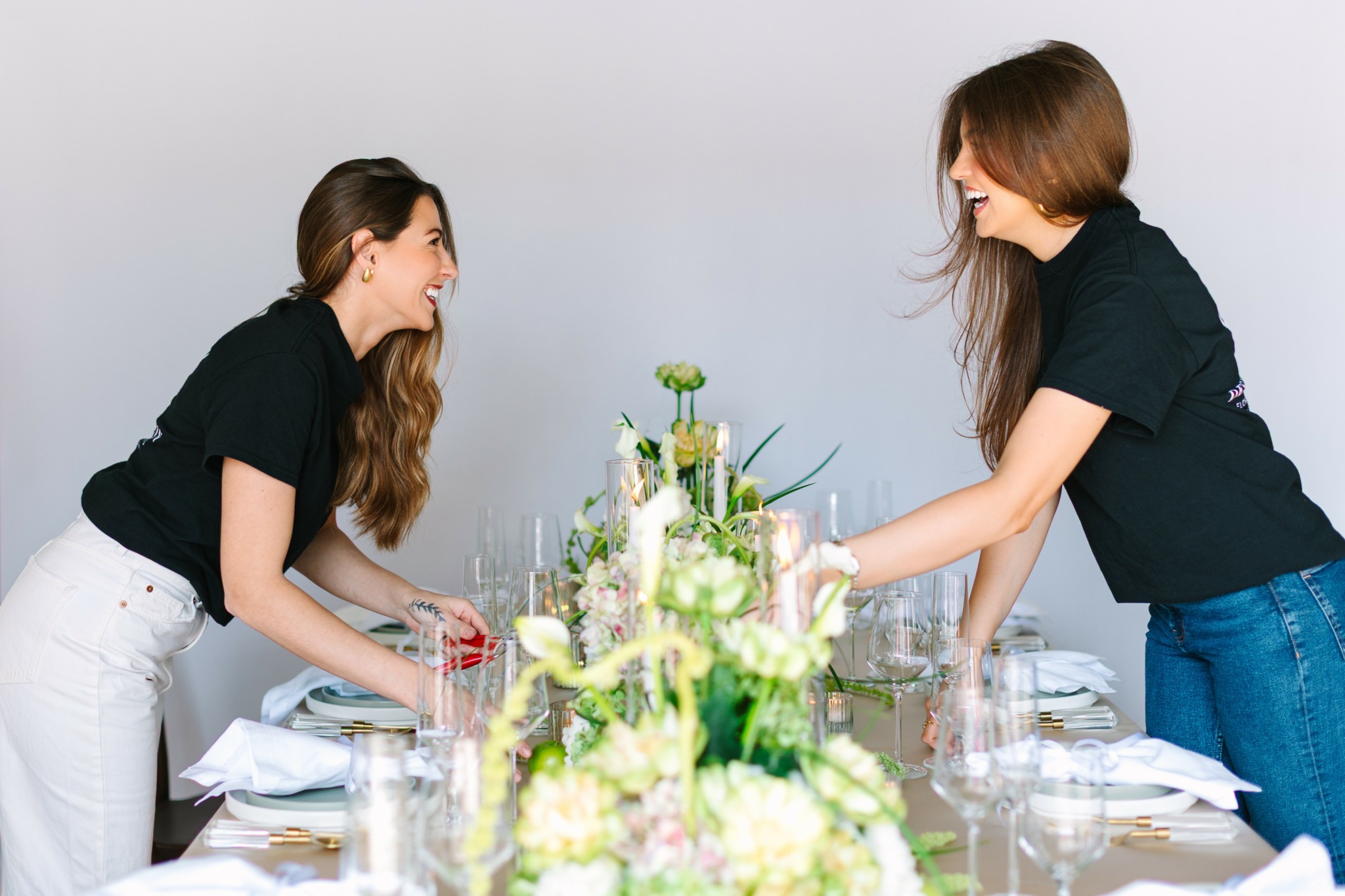 Two women with long hair smiling and talking at a table with flowers and glassware.