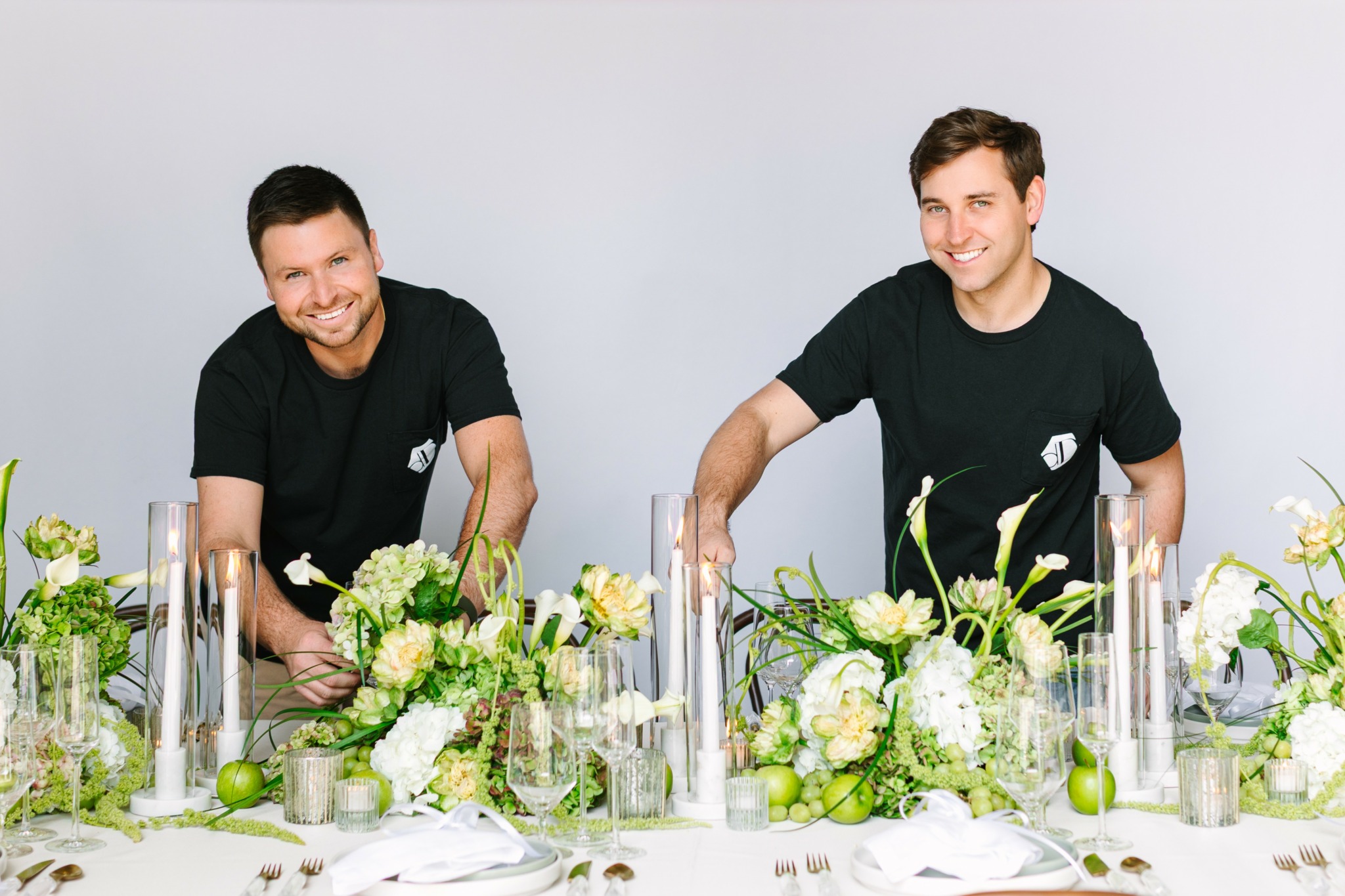 Two men smiling at a decorated table with white and green floral arrangements and candles.
