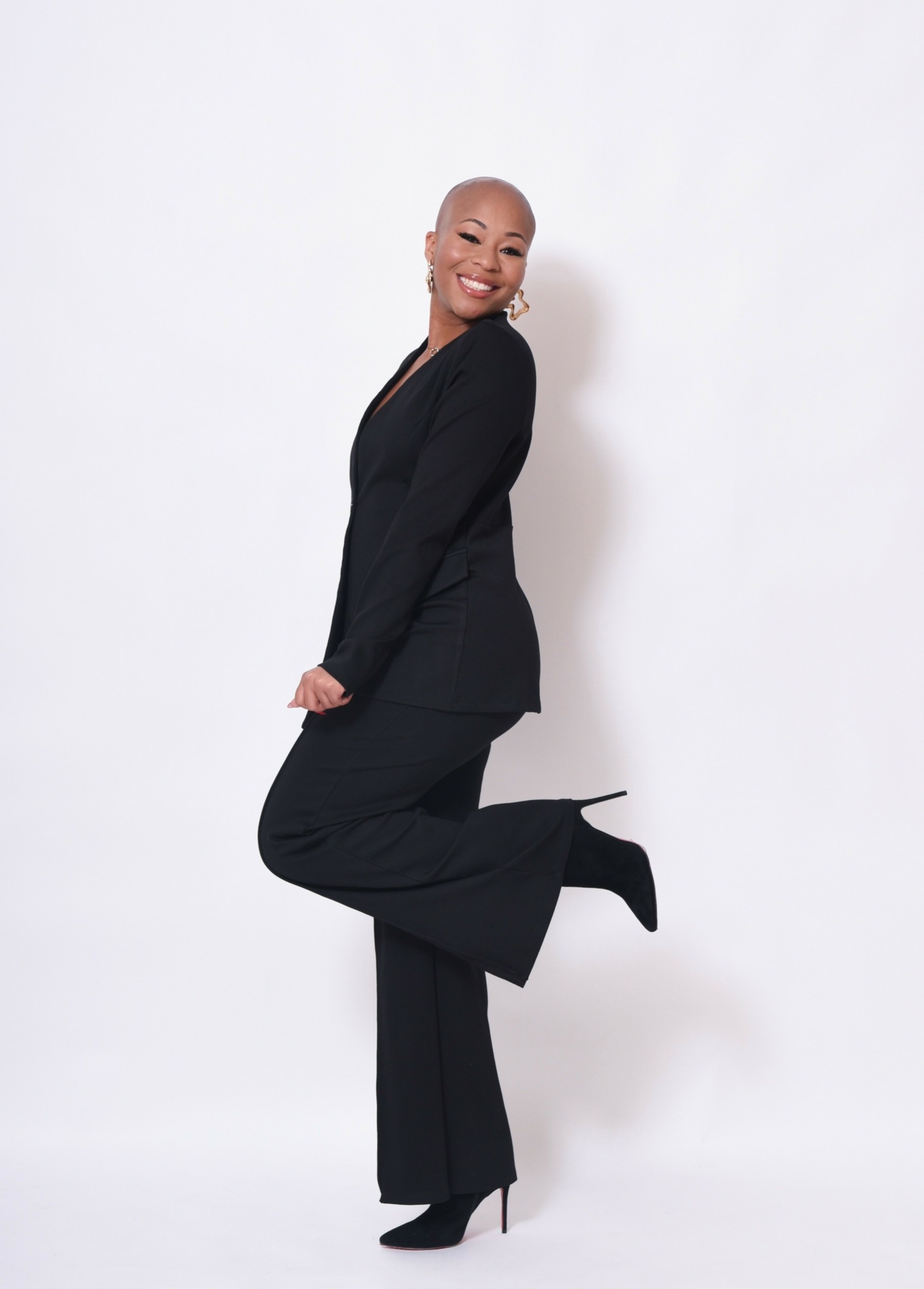 Woman in black suit smiling, standing on one leg with the other leg bent and foot raised.