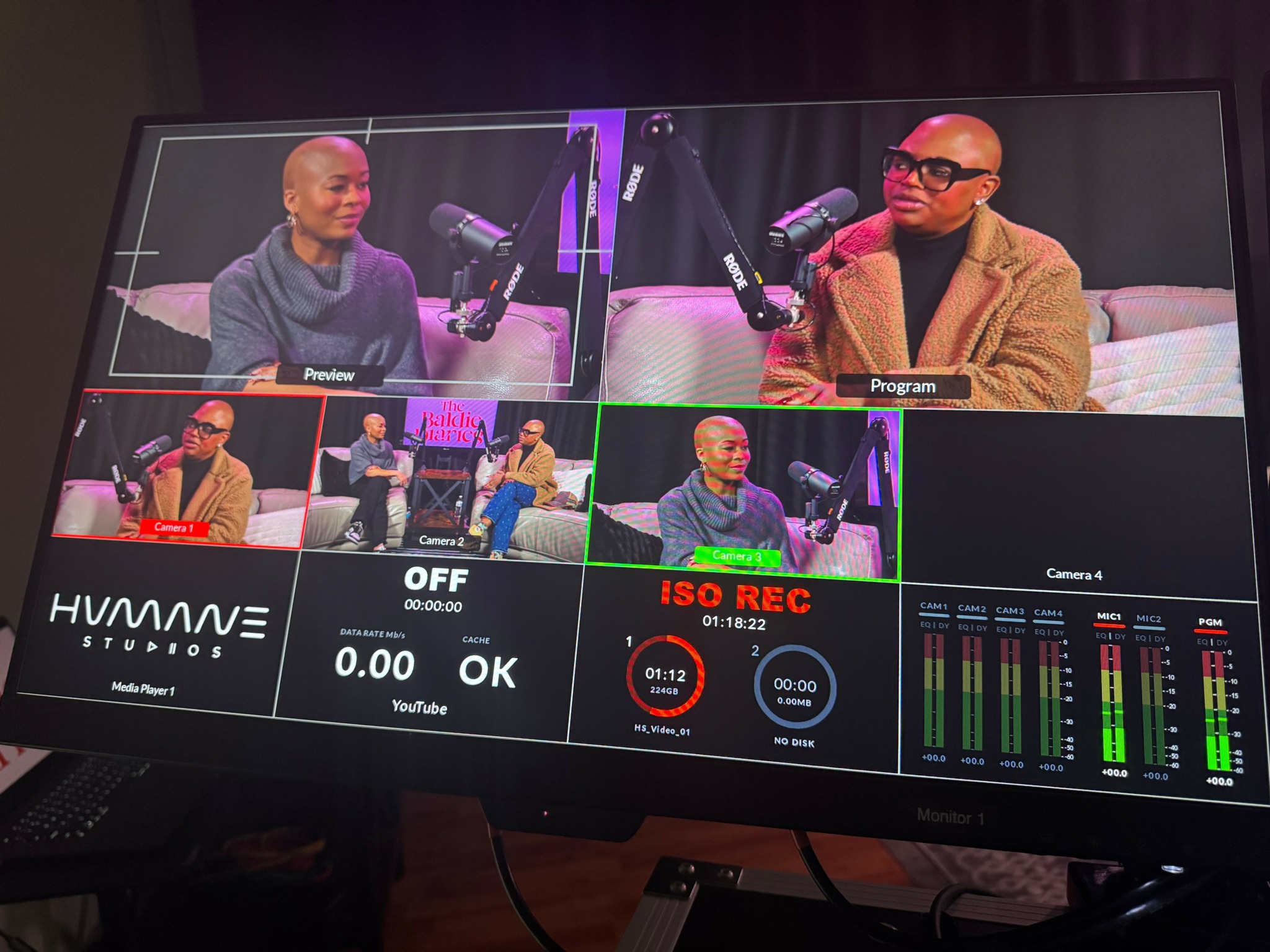 Television screen showing a video recording session with two people speaking into microphones, multiple smaller screens, and audio levels.