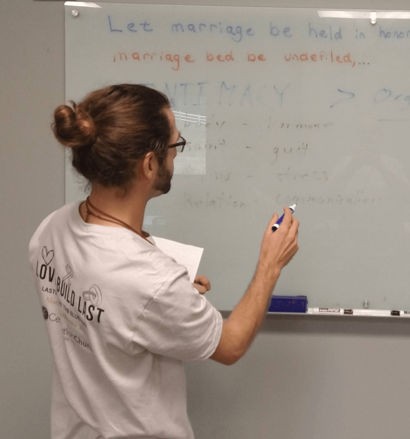 Person with glasses and bun writing on a whiteboard in a classroom or meeting room.