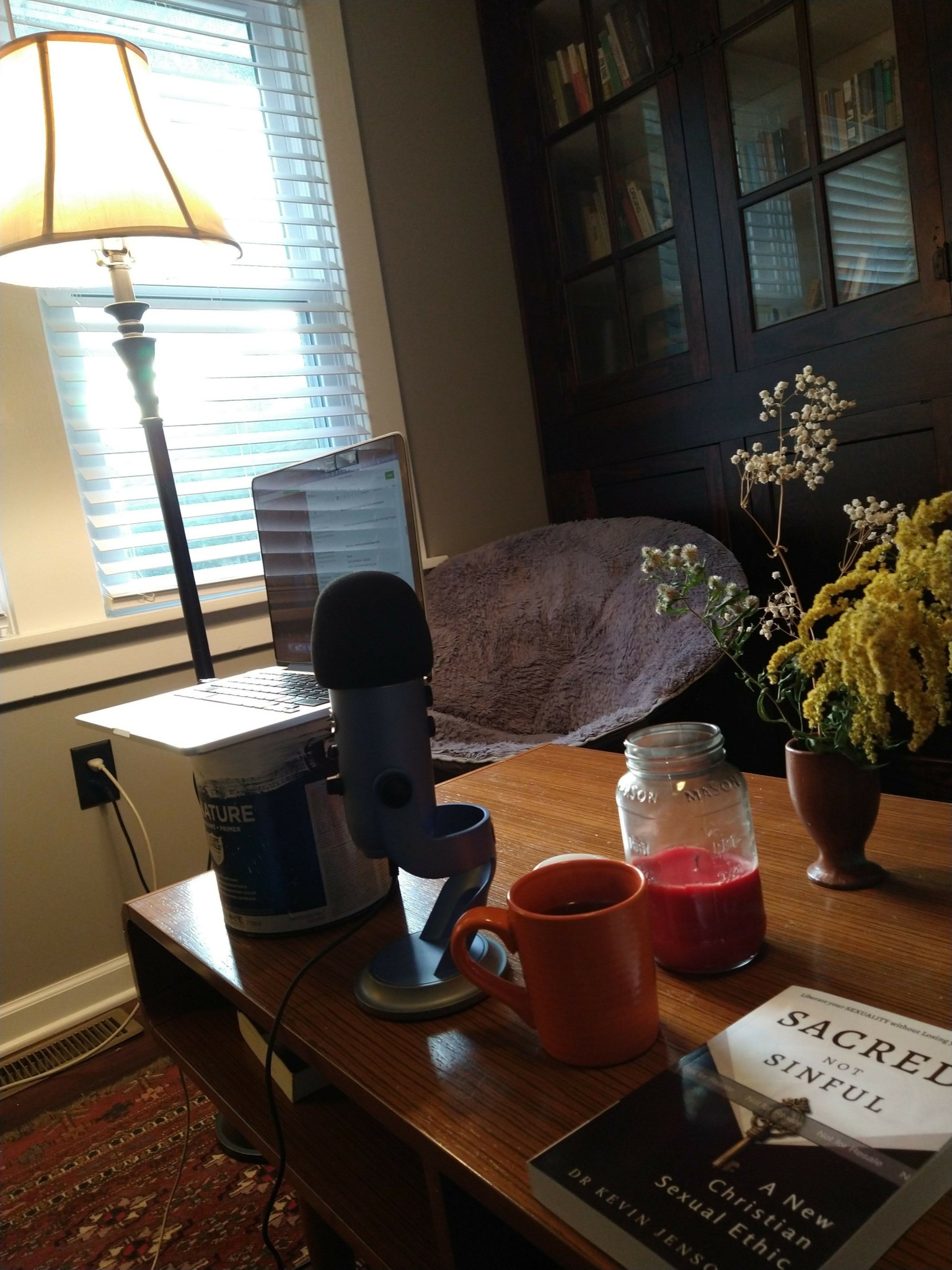 Desk with flowers, microphone, books, and a lamp near a window with blinds.