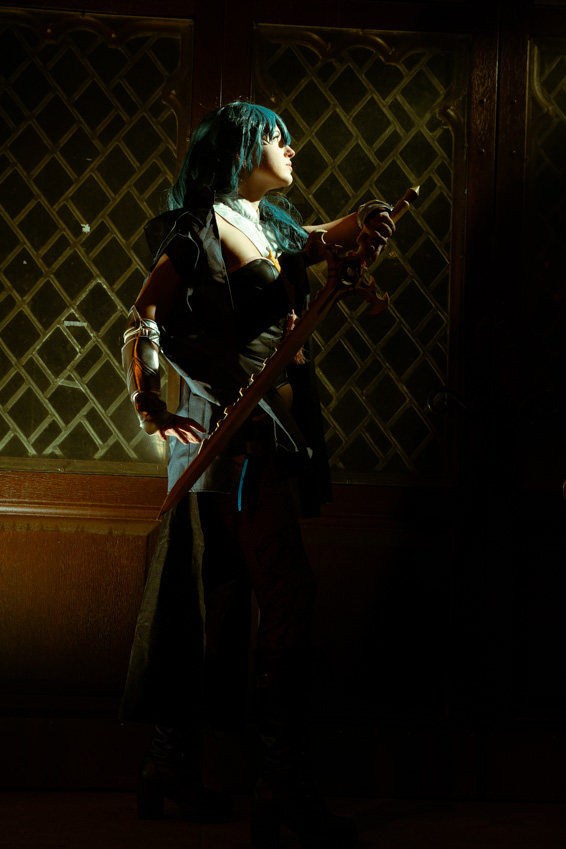 Person with long dark hair and glasses holding a sword, standing in front of a patterned wall, illuminated by a spotlight.
