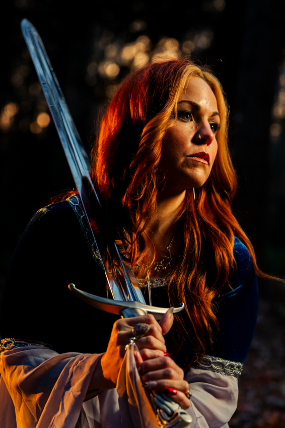 Woman with long red hair holding a sword, outdoors at night with blurred lights in background.