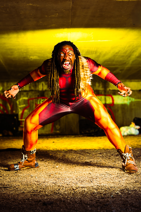 Person with long hair in a red outfit crouches with arms outstretched, shouting intensely on stage with yellow lighting.