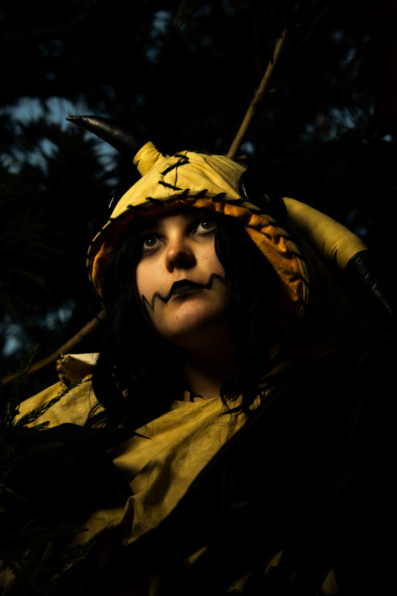 Person in yellow costume with a hat, dark background, serious expression, makeup, and dark hair.