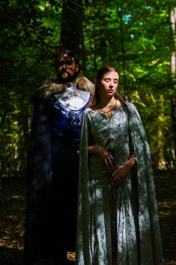 Man and woman in medieval-style costumes standing in a forest with green foliage.