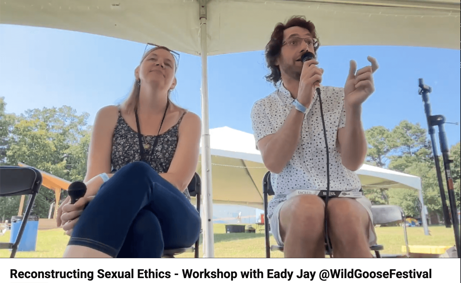 Two people seated outdoors under a tent, one speaking into a microphone, the other listening with a microphone in hand.