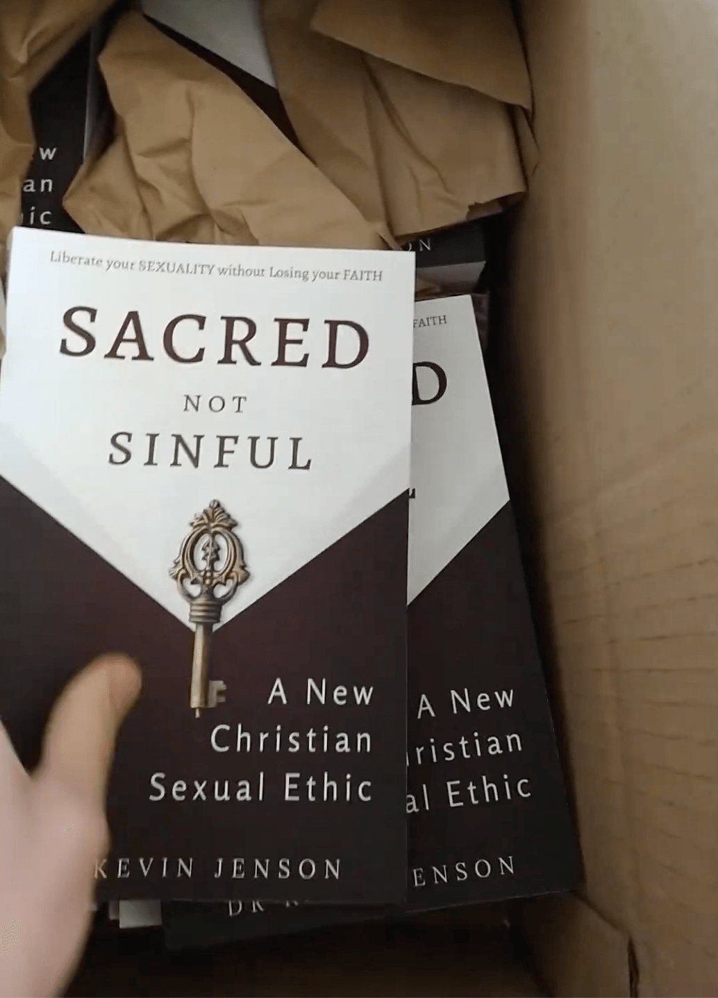 Two copies of a book titled 'Sacred Not Sinful' inside a cardboard box, partially covered by brown paper.